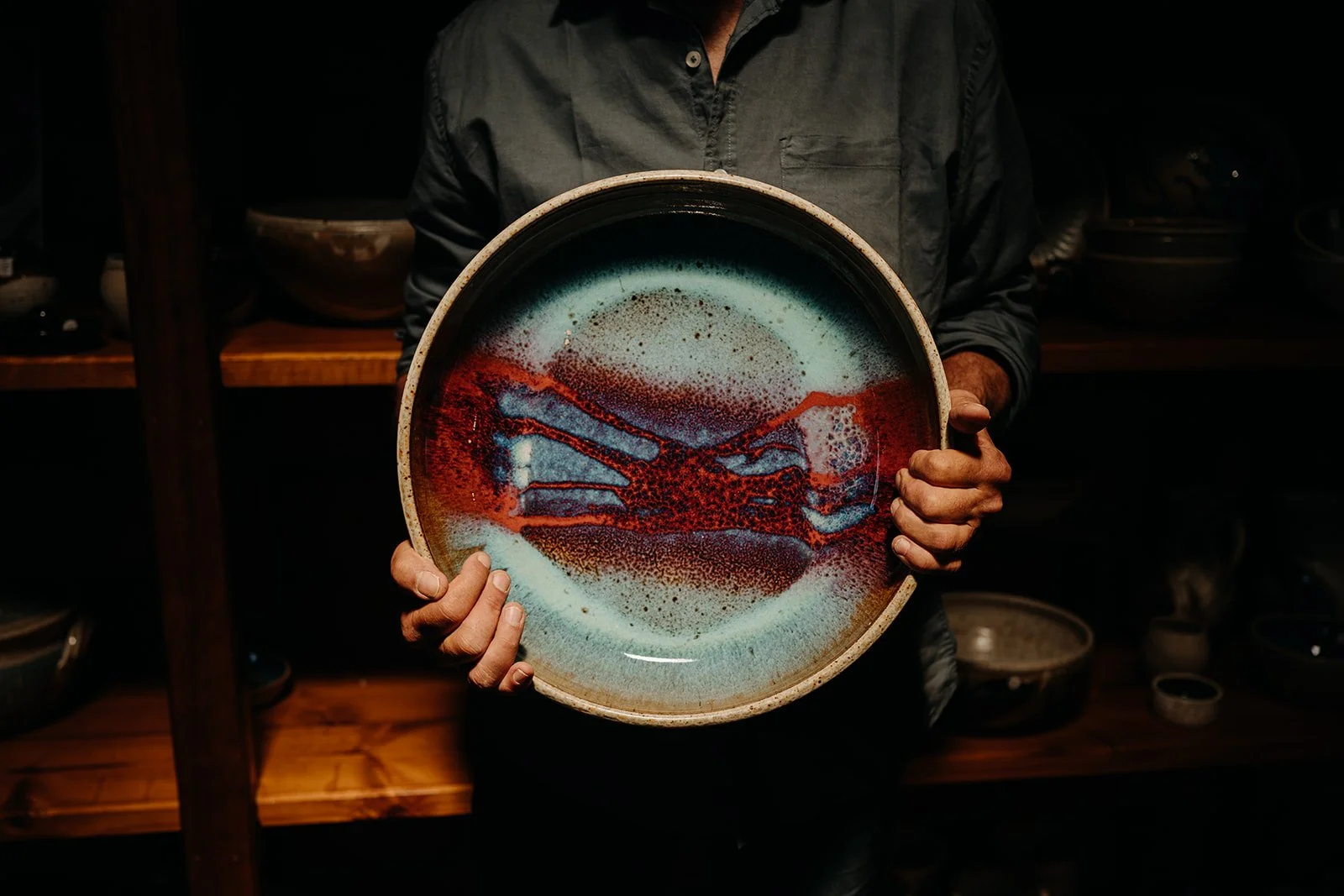 Person holding a decorative ceramic plate with an abstract design in red, blue, and beige colors. The background shows wooden shelves with other pottery items.