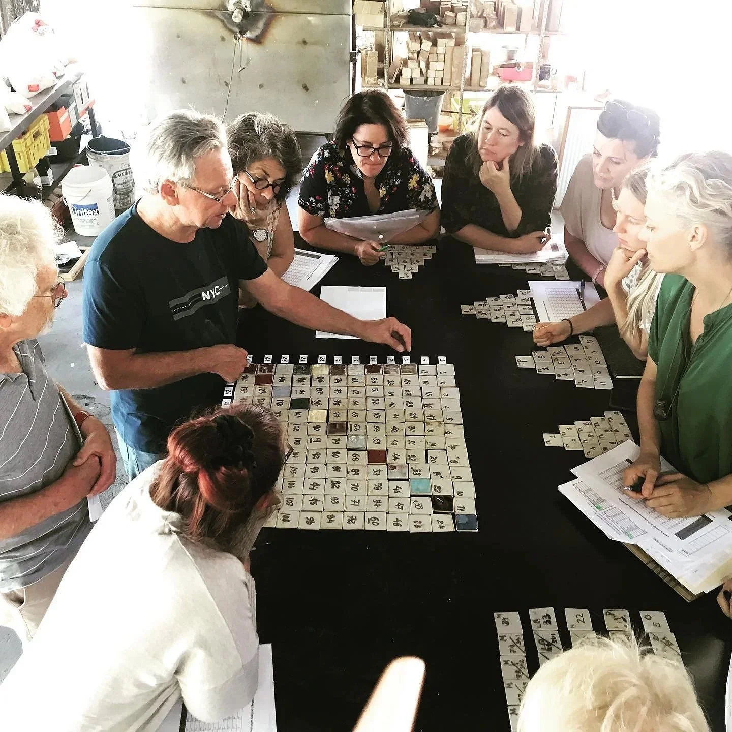 A group of people gathered around a table playing a game with tiles that have numbers on them, in a workshop or studio setting.