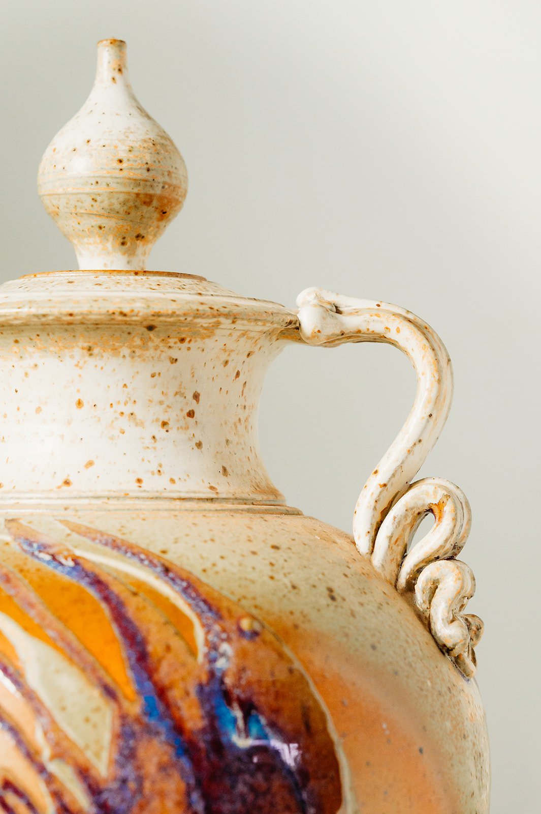 Close-up of a ceramic vase with a textured beige and orange painted design, featuring a curvy handle and a decorative lid with a knob, against a plain light background.