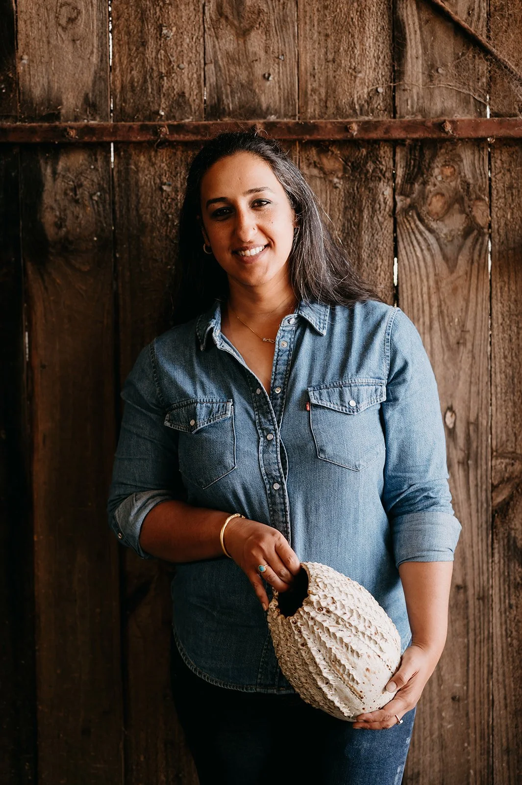 A woman with long dark hair wearing a denim shirt, holding a large mushroom, standing in front of a wooden wall.