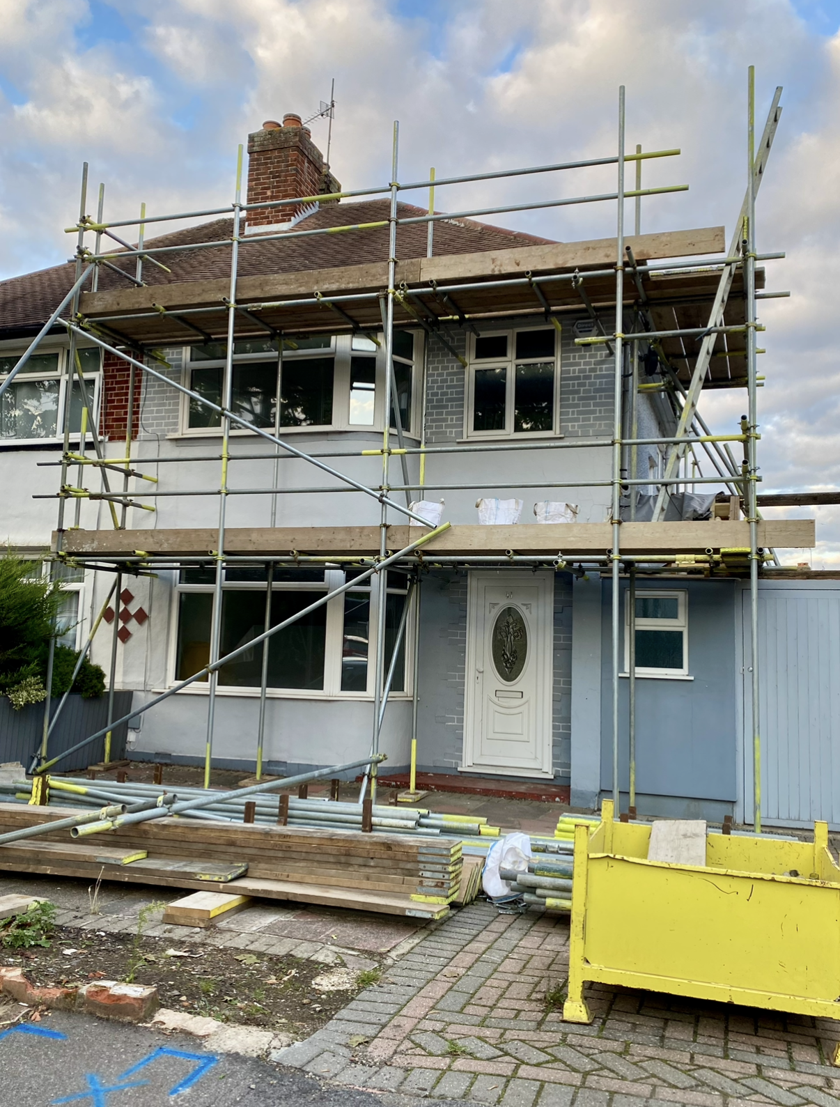 A house under renovation with scaffolding around it. The house has a white lower section, a gray brick section above the door, three windows on the upper level, and construction materials in front, including wooden planks, metal poles, and a yellow container.