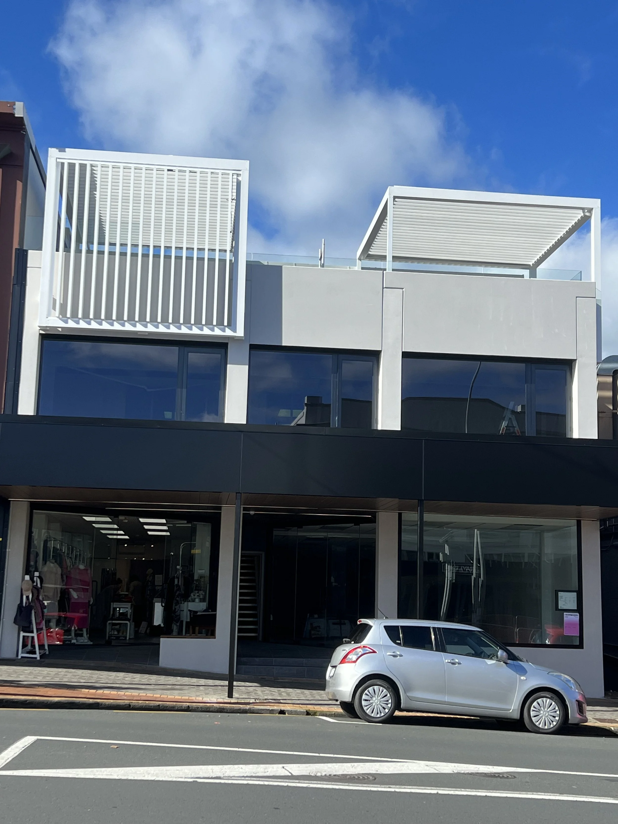 Modern two-story building with large glass windows and white architectural elements, including a patio with slatted rooftops. A silver car is parked in front on a city street under a blue sky with clouds.