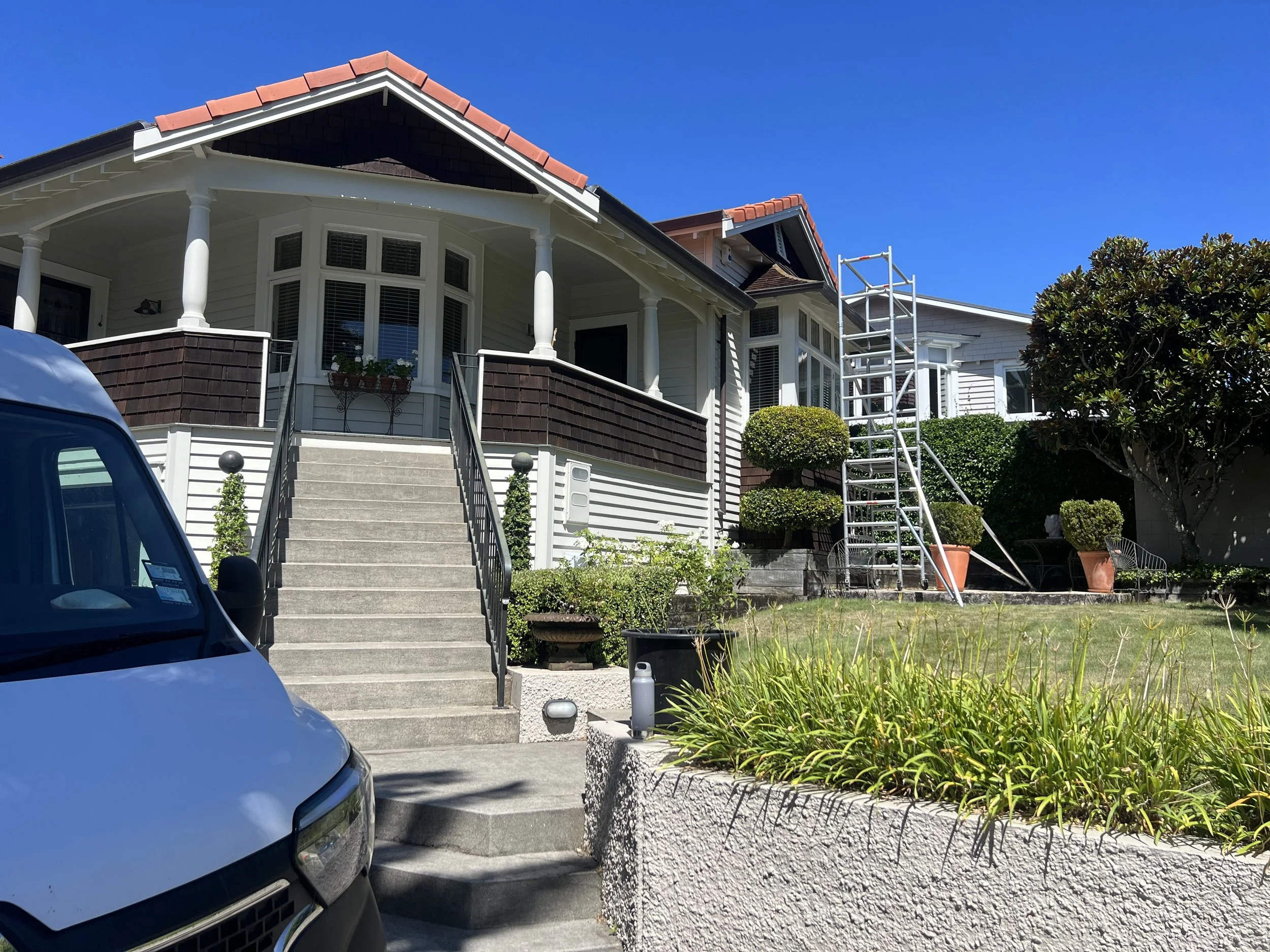 Front view of a large house with a staircase leading up to the front door, surrounded by greenery and a driveway with parked cars, under a clear blue sky.