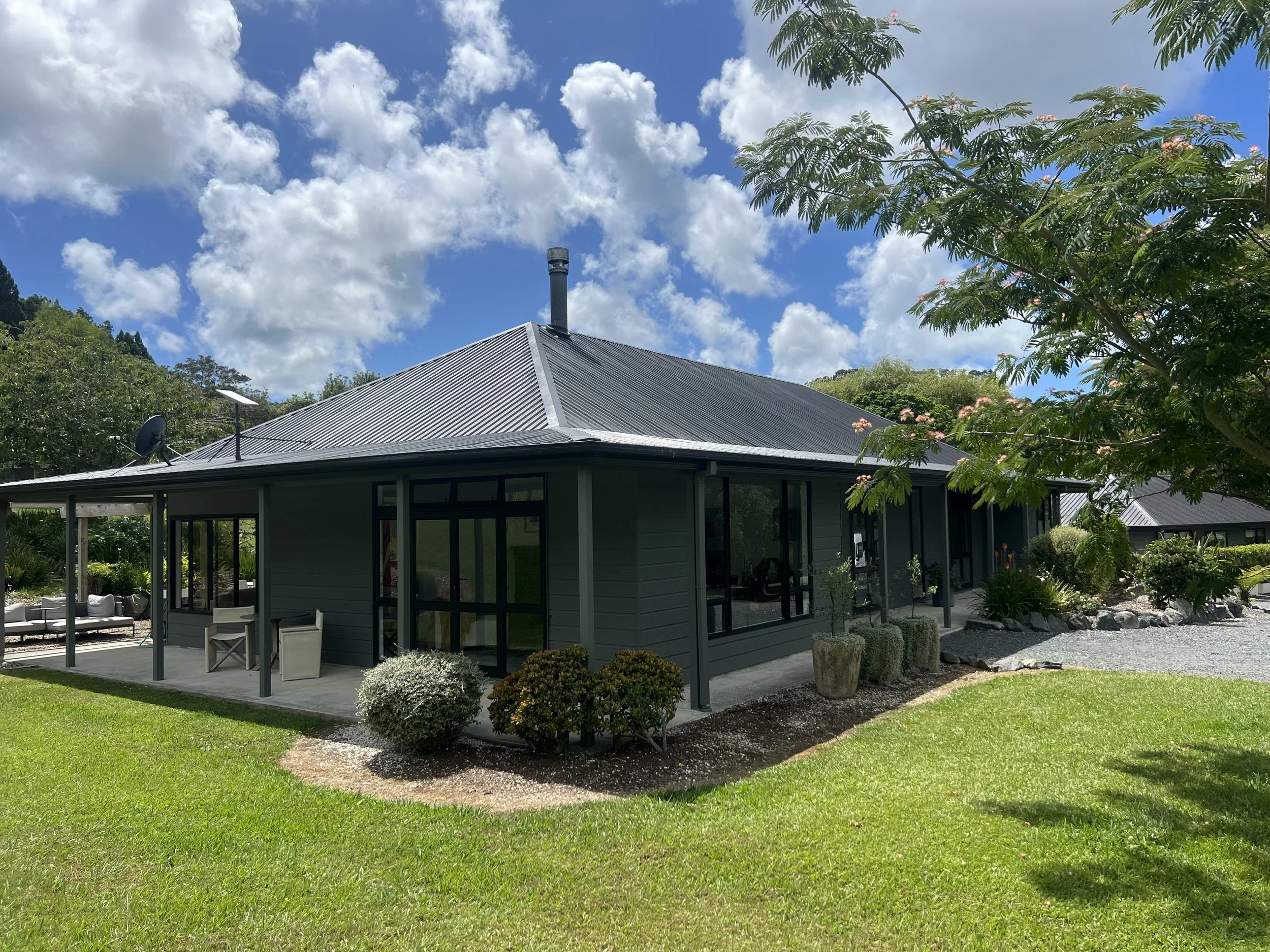 A house with a dark gray, metal roof, surrounded by green grass and shrubbery, with trees and a blue sky with white clouds above.