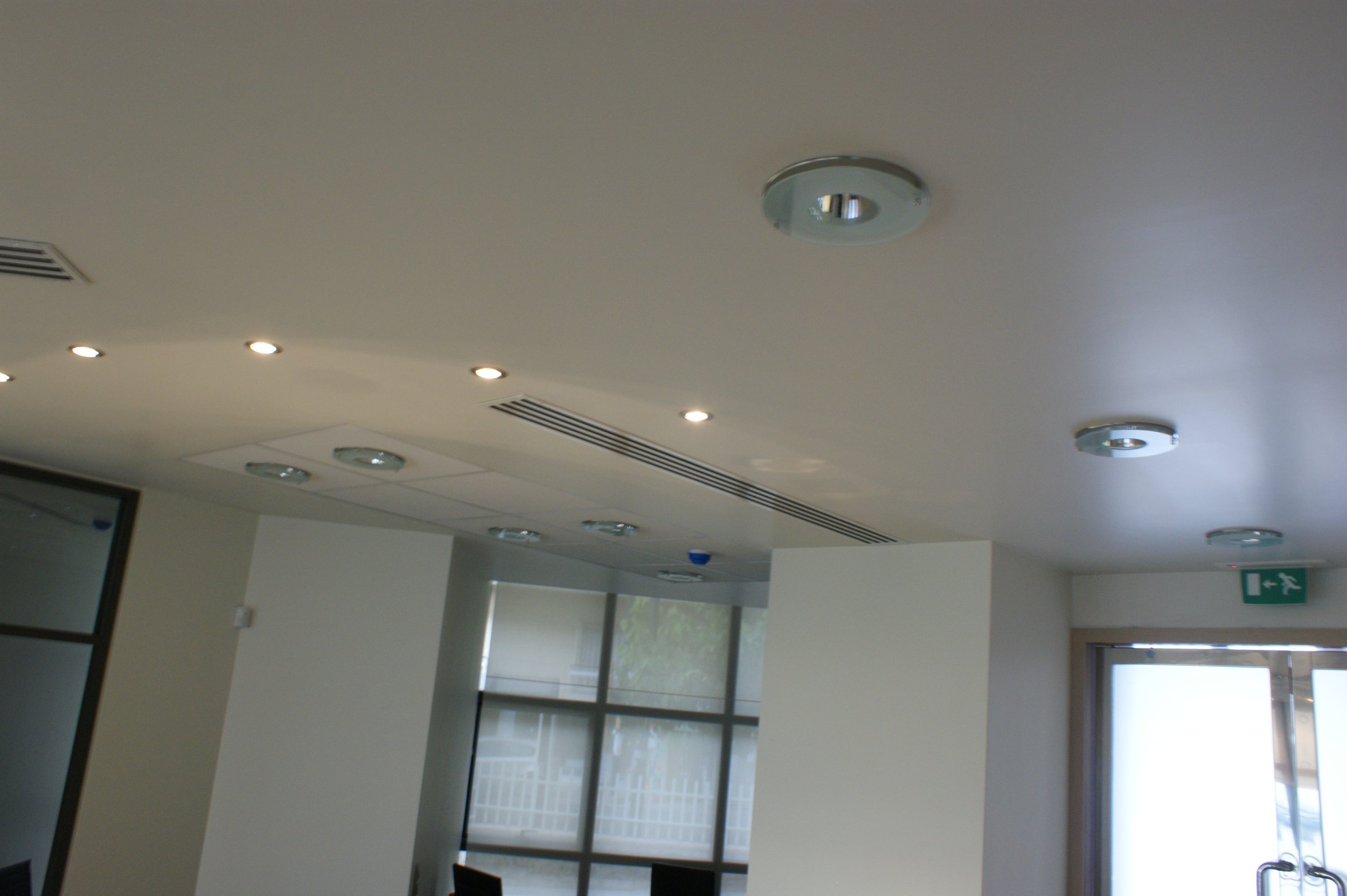Empty indoor room with a white ceiling, ceiling lights, vents, large windows, and a door with an exit sign.
