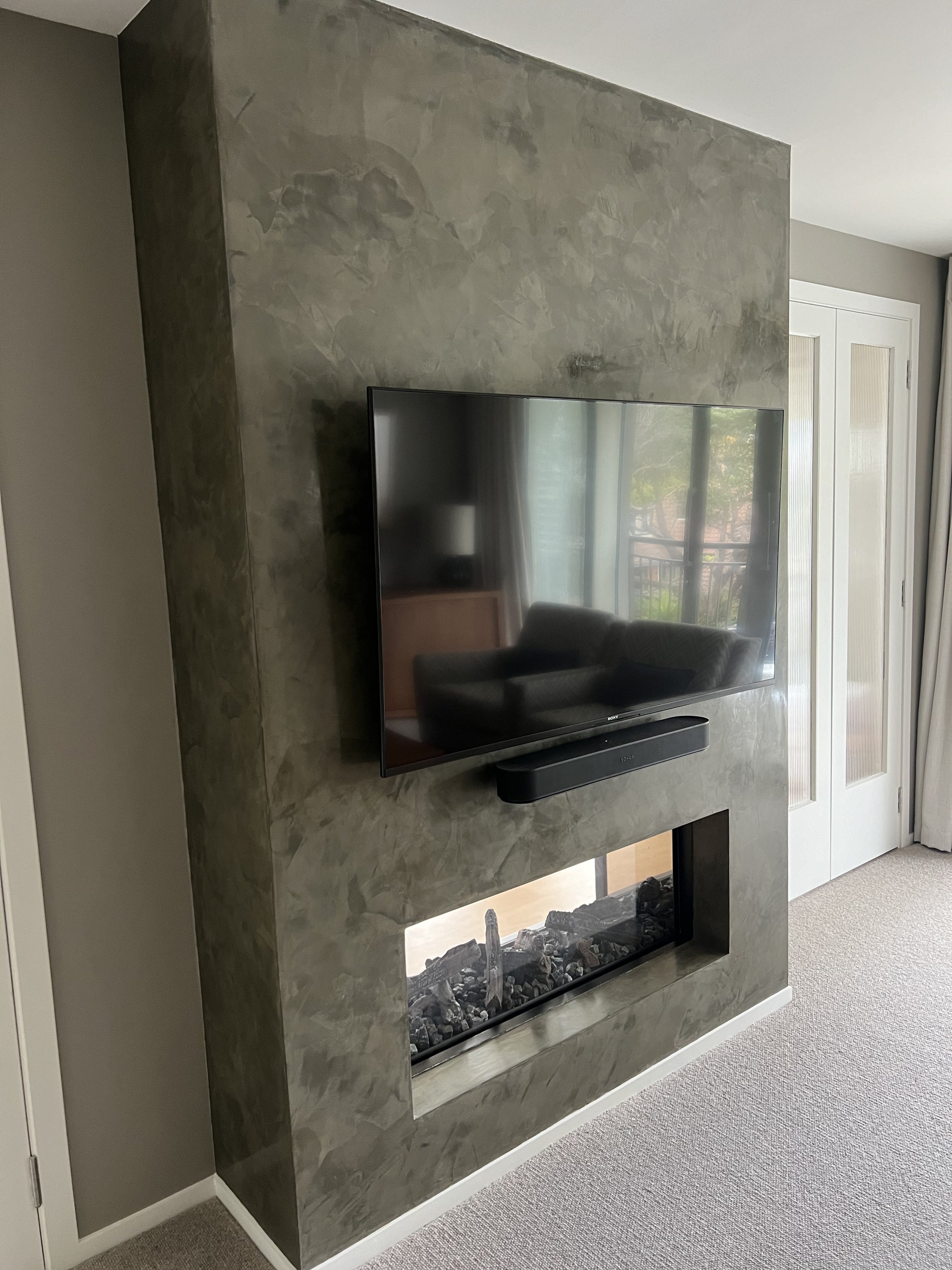A modern fireplace with a stone finish, a flat-screen TV mounted above it, and a soundbar beneath the TV. The fireplace has black rocks inside and is located in a room with beige carpet and a white door.