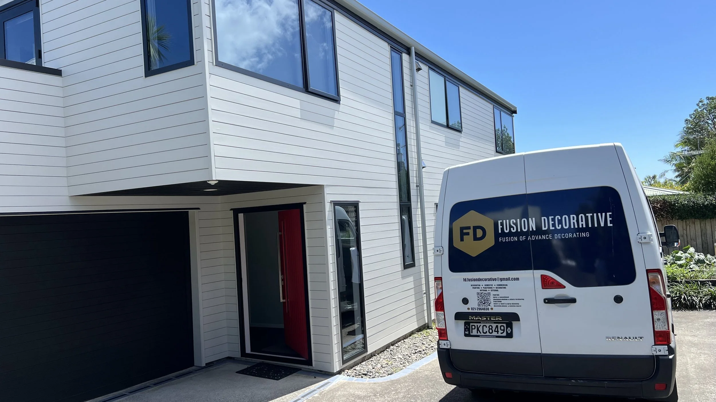 Modern white building with large windows and a black garage door, parked white van with company logo, blue sky, green trees.