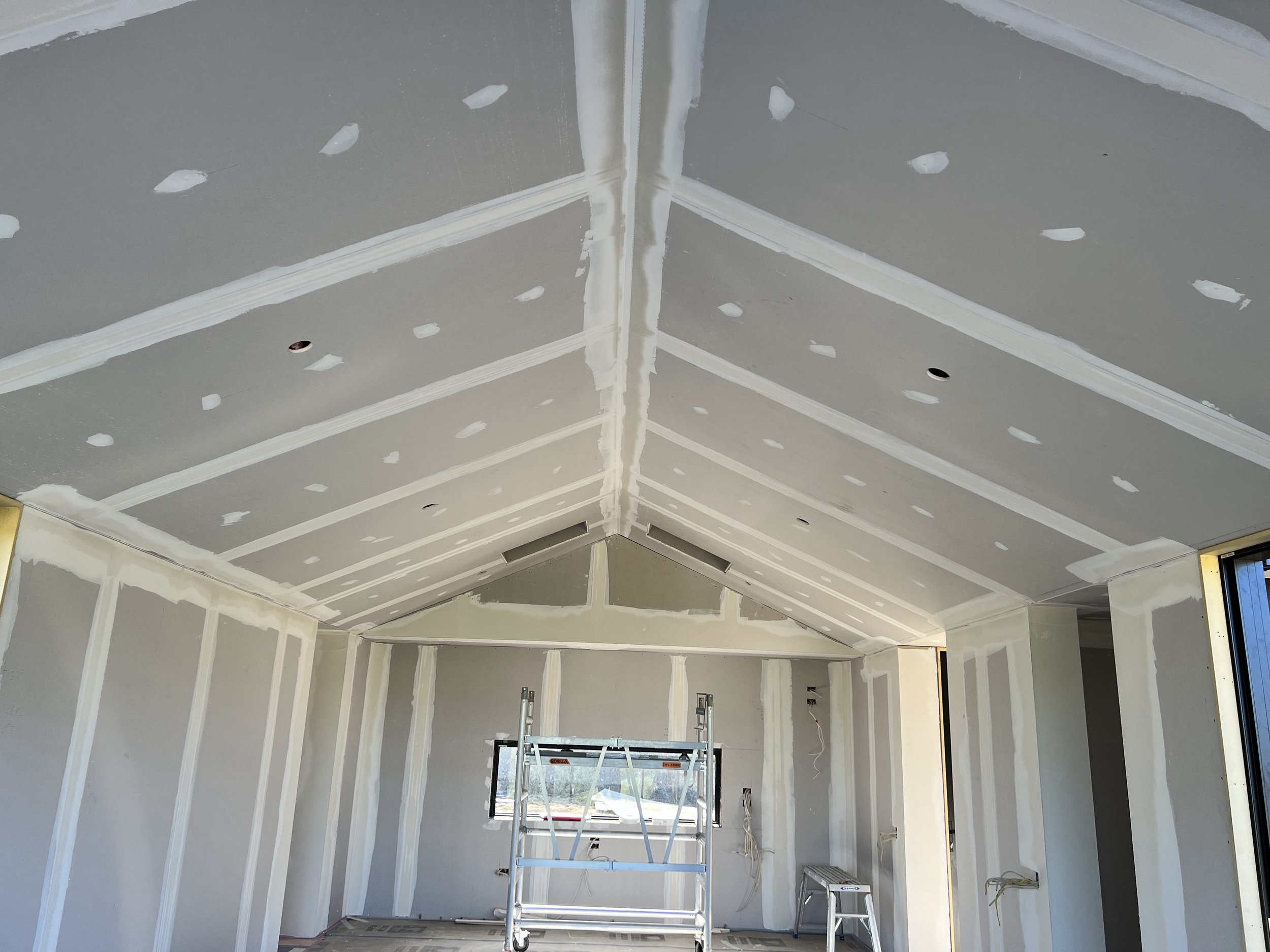 Interior of a room under construction with drywall installed and joint compound applied, scaffolding in the center, and wiring hanging from the ceiling.