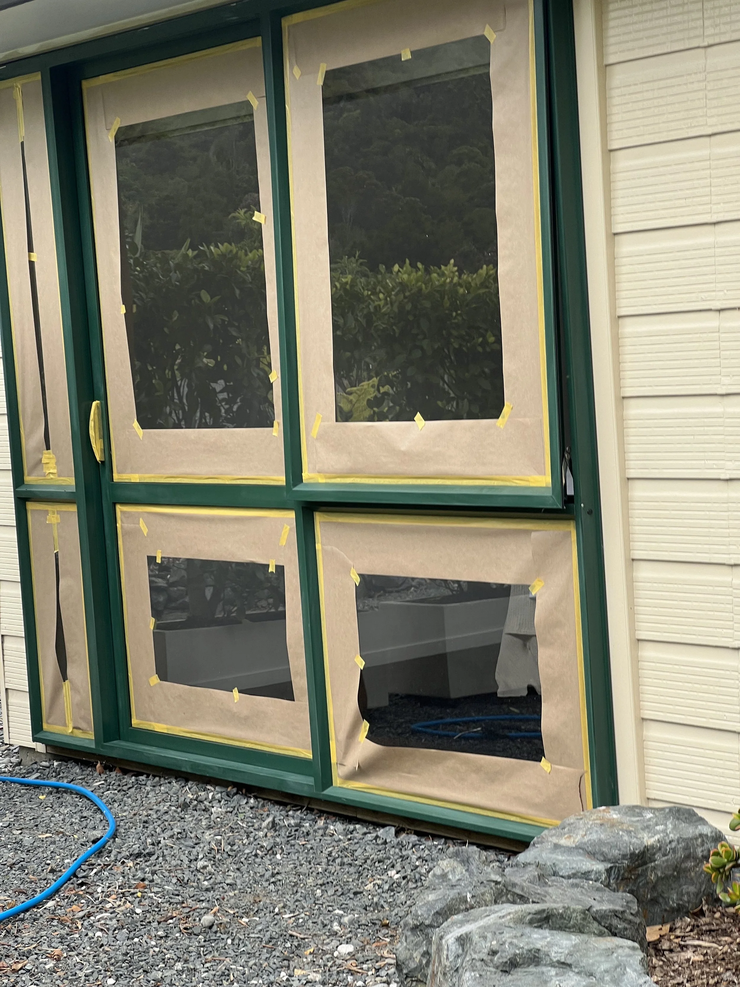 A large green-framed window or door being installed on a house, covered with masking paper taped around the edges, with rocks, gravel, and a blue hose on the ground outside.