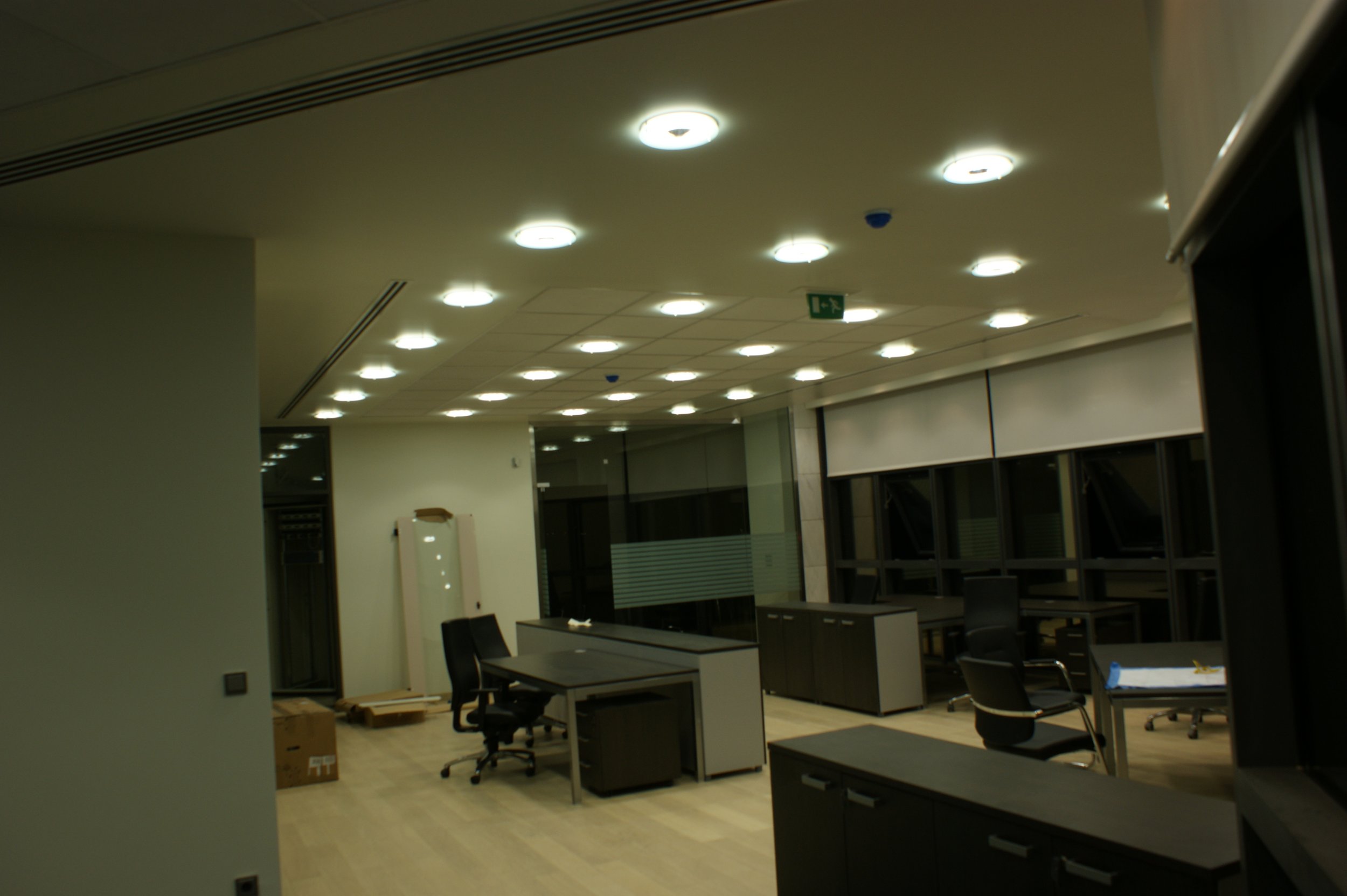 An interior office space with ceiling lights, office desks, chairs, and some boxes on the floor, viewed from a partially open doorway.