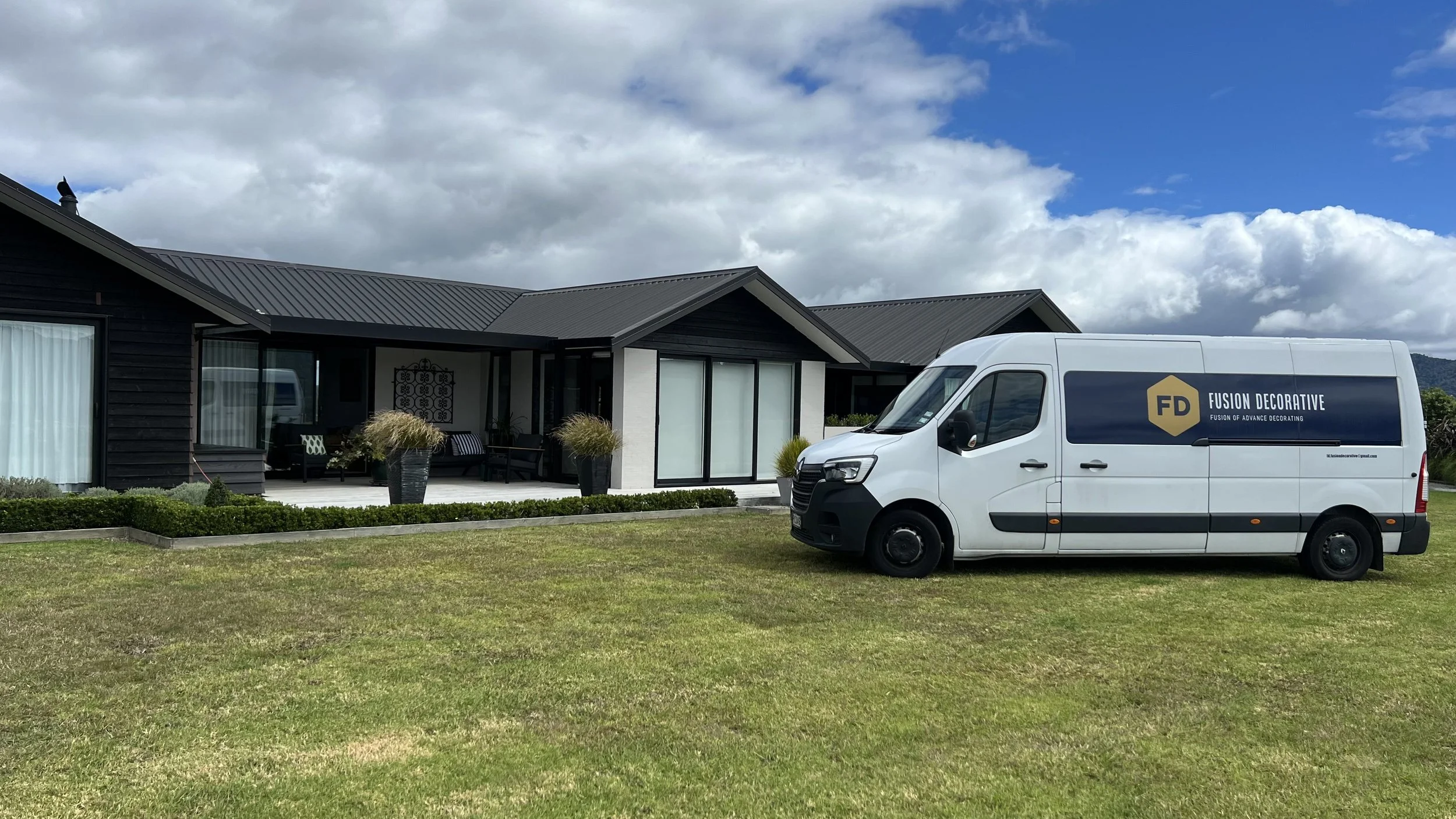 A modern house with a gray metal roof and black exterior walls, with a porch area featuring outdoor furniture and large potted plants. A white service van with the logo 'Fusion Decorative' is parked on the lawn in front of the house under a partly cl
