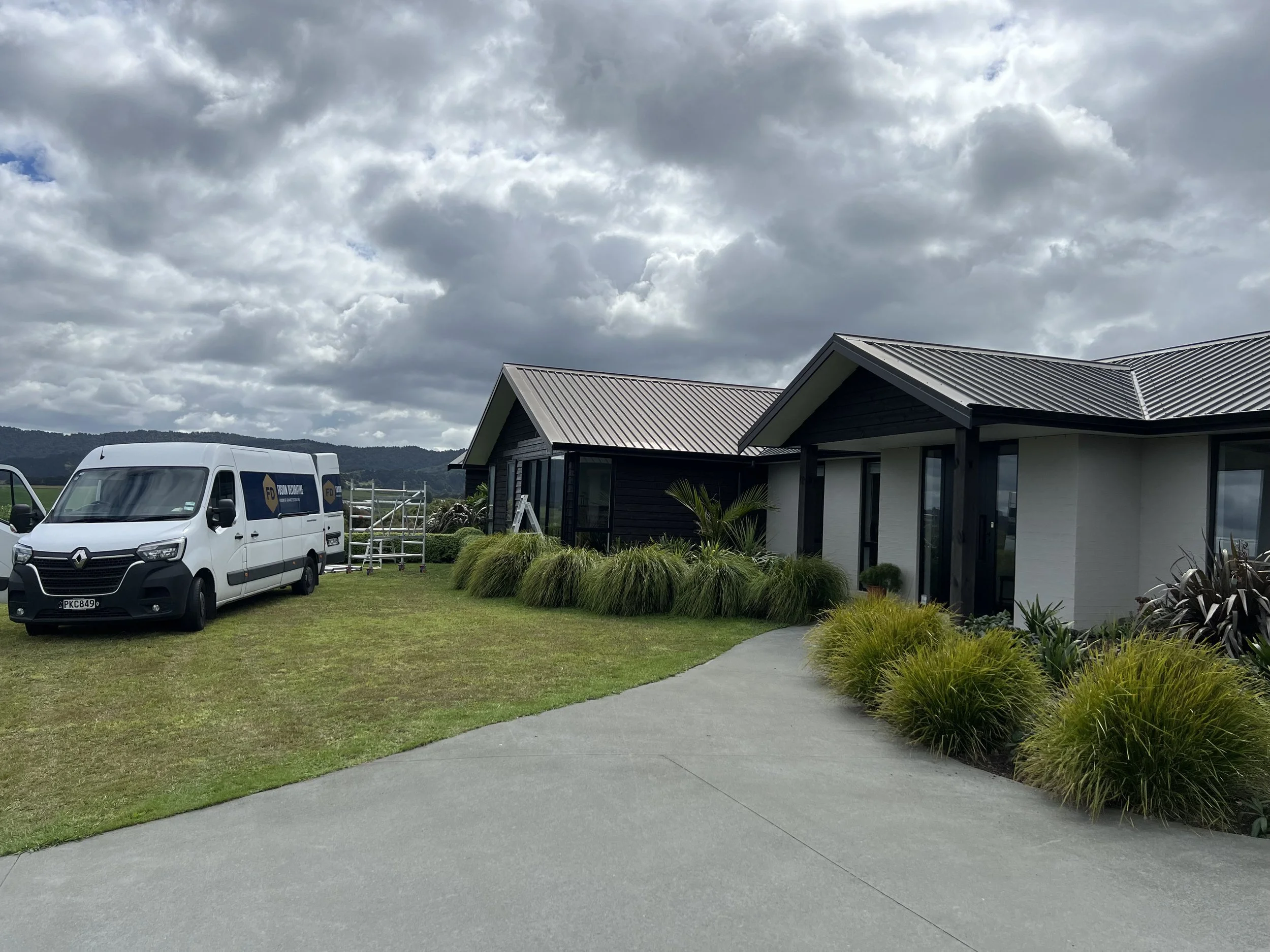 Exterior view of a modern house with black siding and a metal roof, surrounded by greenery, with a white van parked nearby and cloudy sky overhead.
