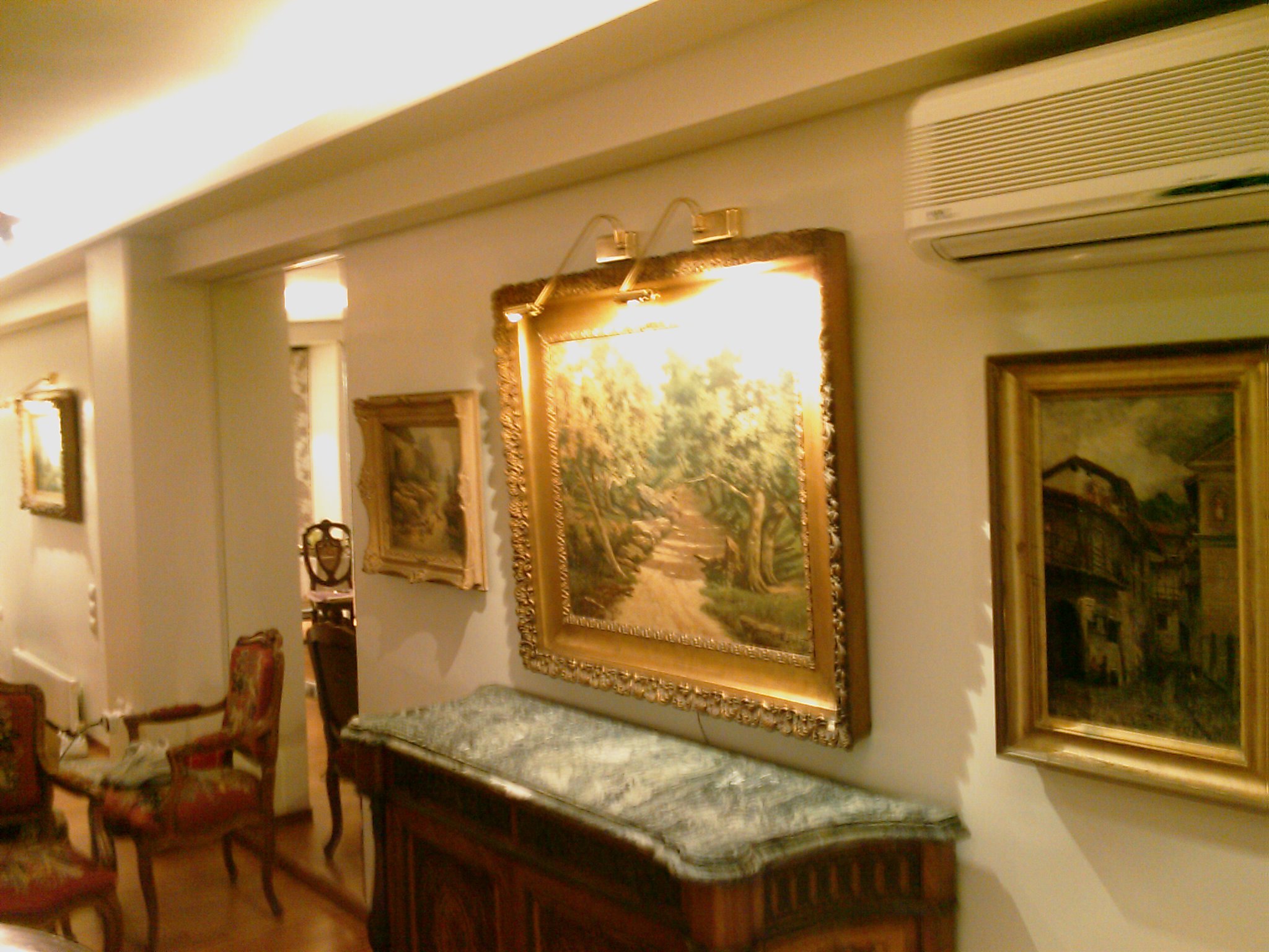 A room with classical furniture and framed paintings on the wall, including a large landscape painting with a decorative gold frame, baroque-style chairs with floral upholstery, and a marble-top sideboard.