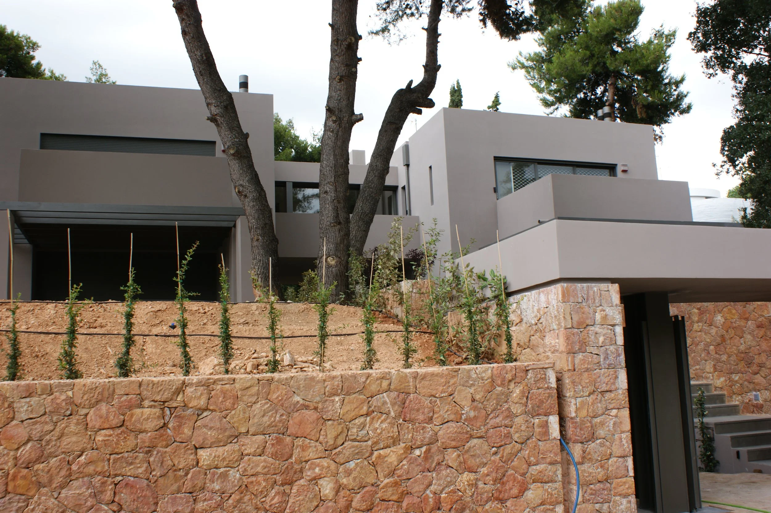 Modern white residential building with multiple levels, large windows, and stone foundation, surrounded by trees and a garden with young plants, under an overcast sky.