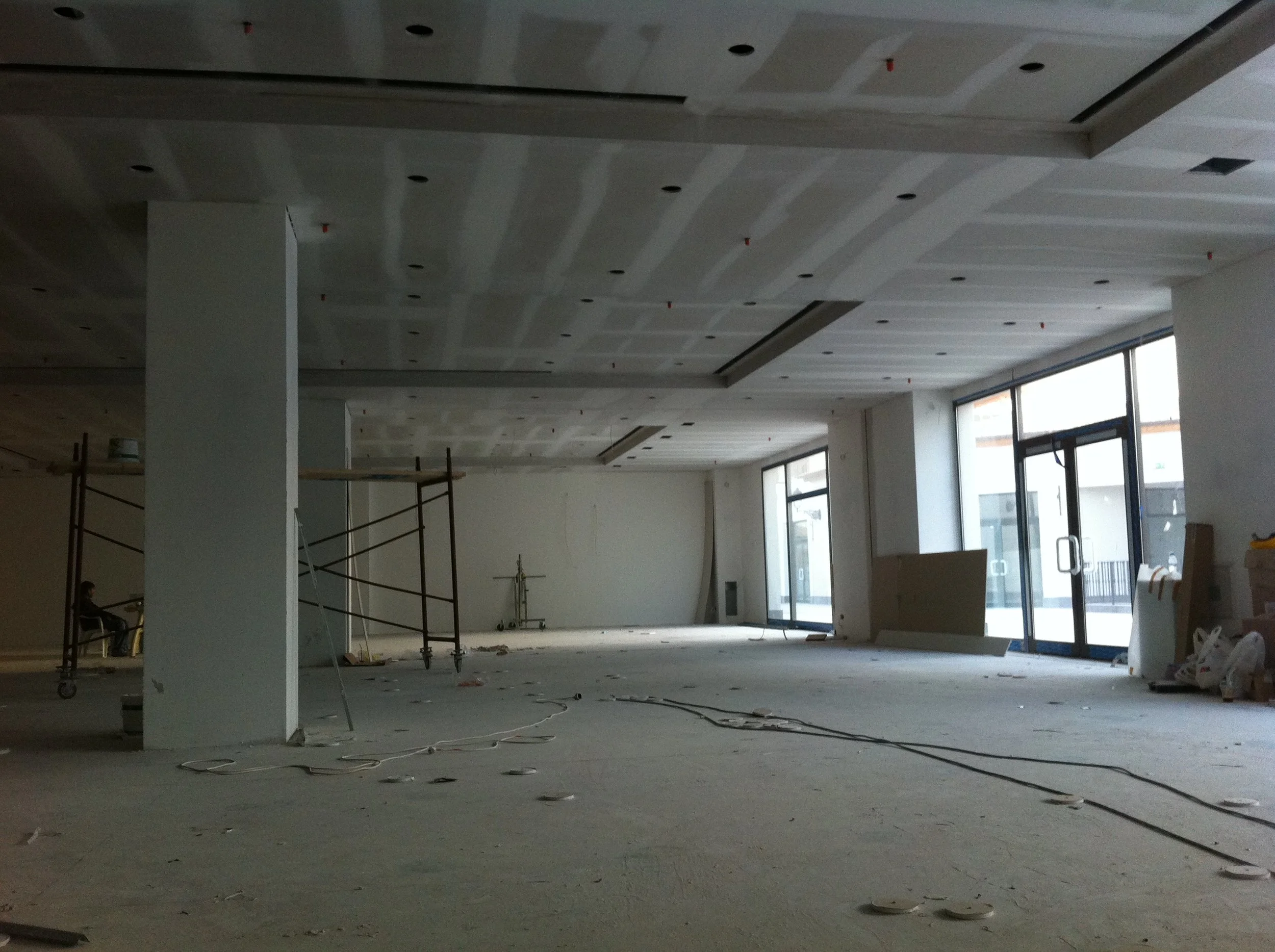 Interior of a building under construction with unfinished floors, exposed wiring, scattered construction materials, scaffolding, and large windows.