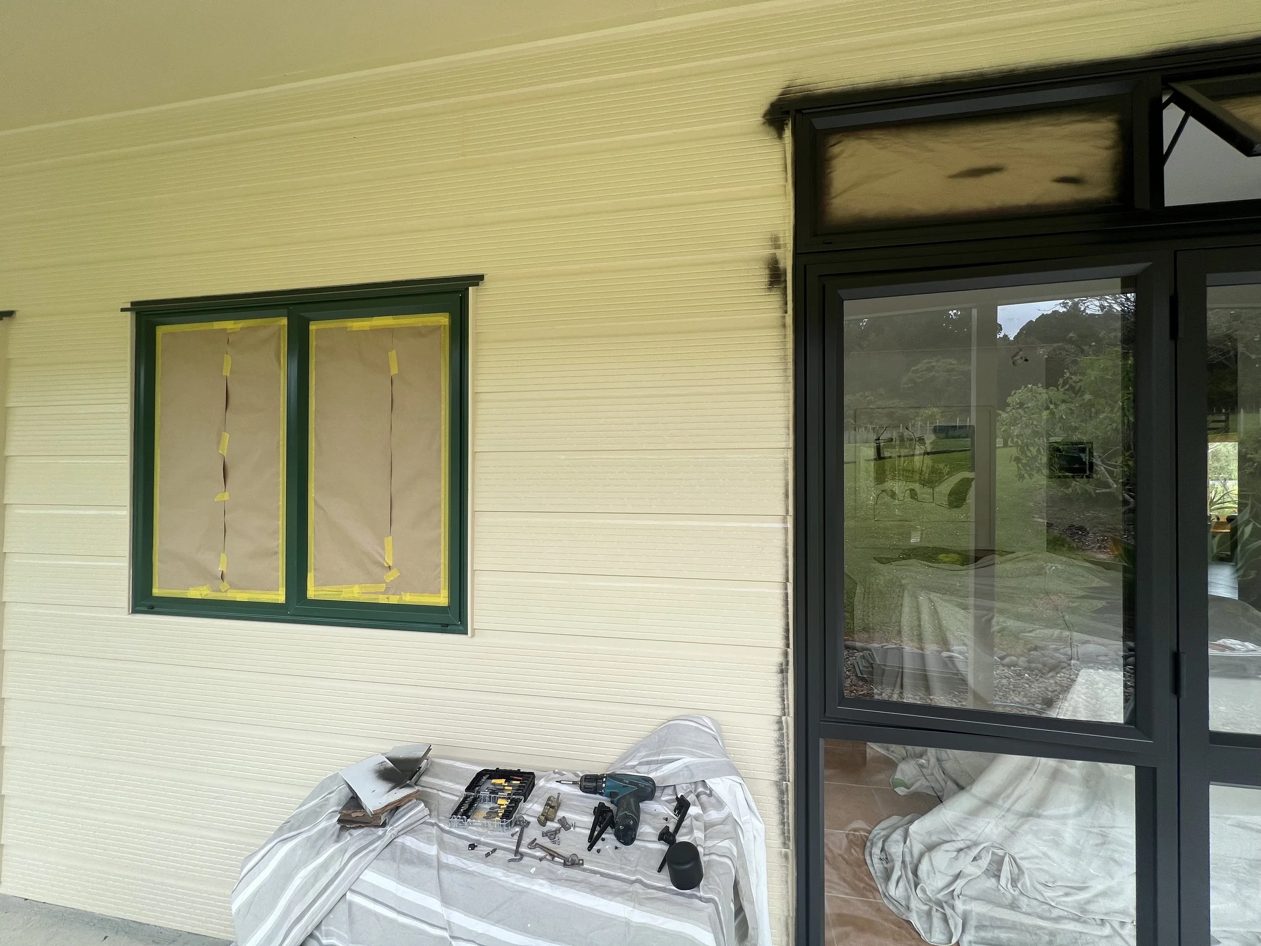 The exterior wall of a house with recently painted or repaired siding, with some tools and materials like drills, screws, and paper covering furniture or the floor inside the house.