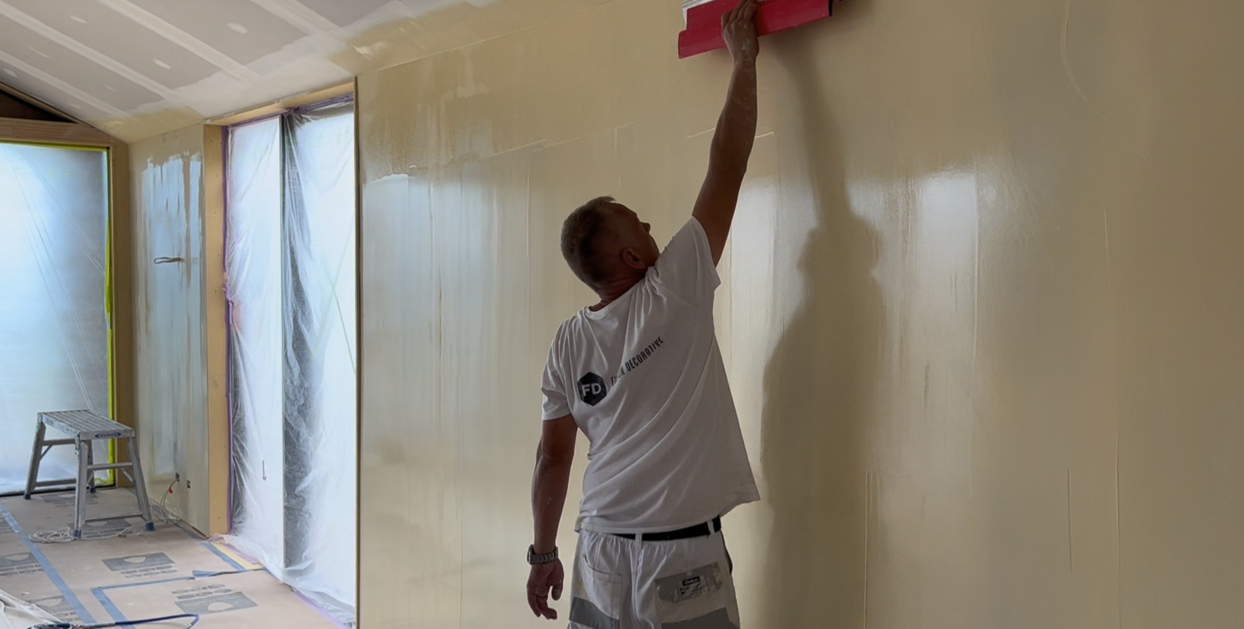 A man skimm coating in a room under construction, with plastic coverings on windows and protective floor covering.