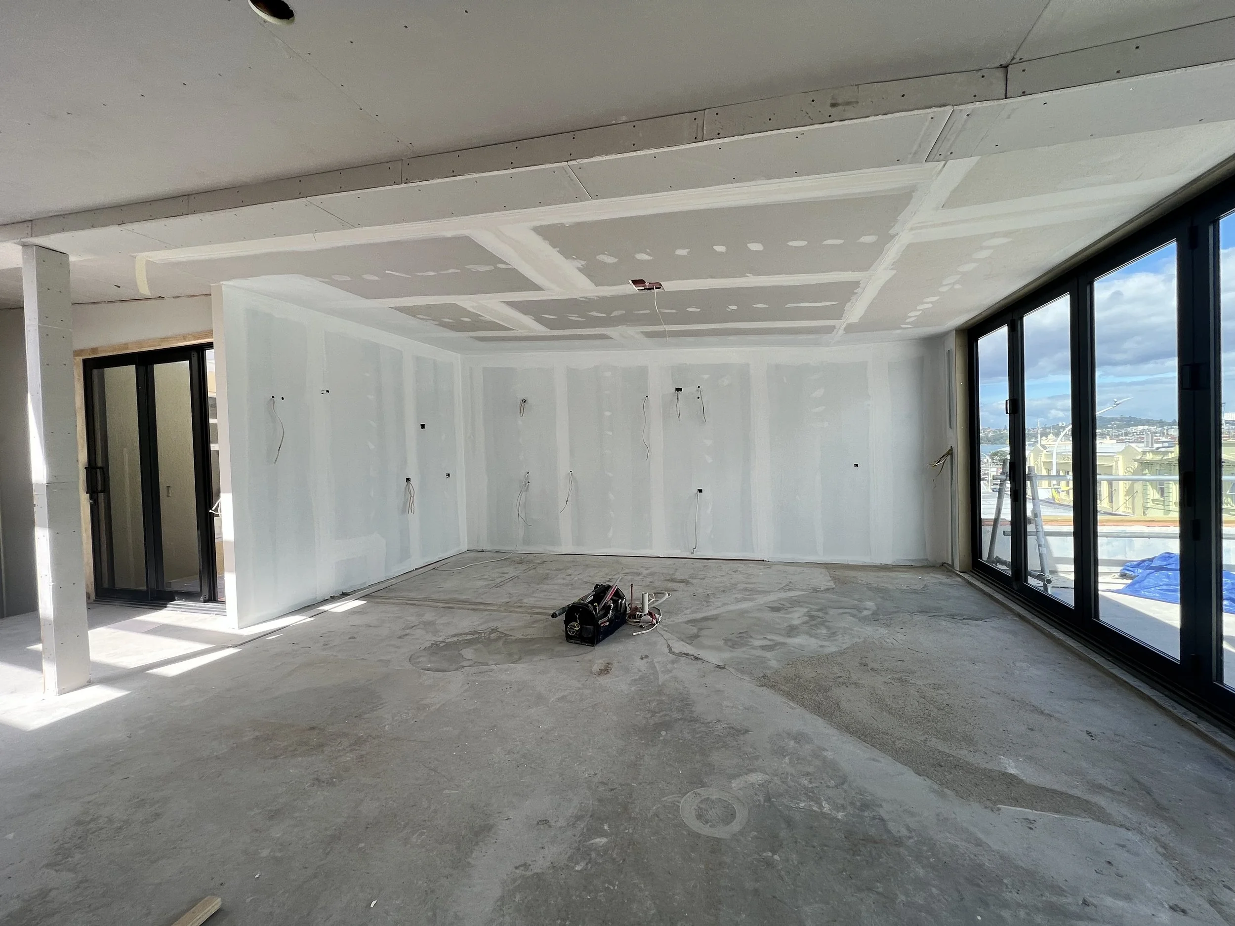 Interior of a building under construction with unfinished drywall on the ceiling and walls, exposed wiring, large sliding glass doors, and a view of a cityscape outside.