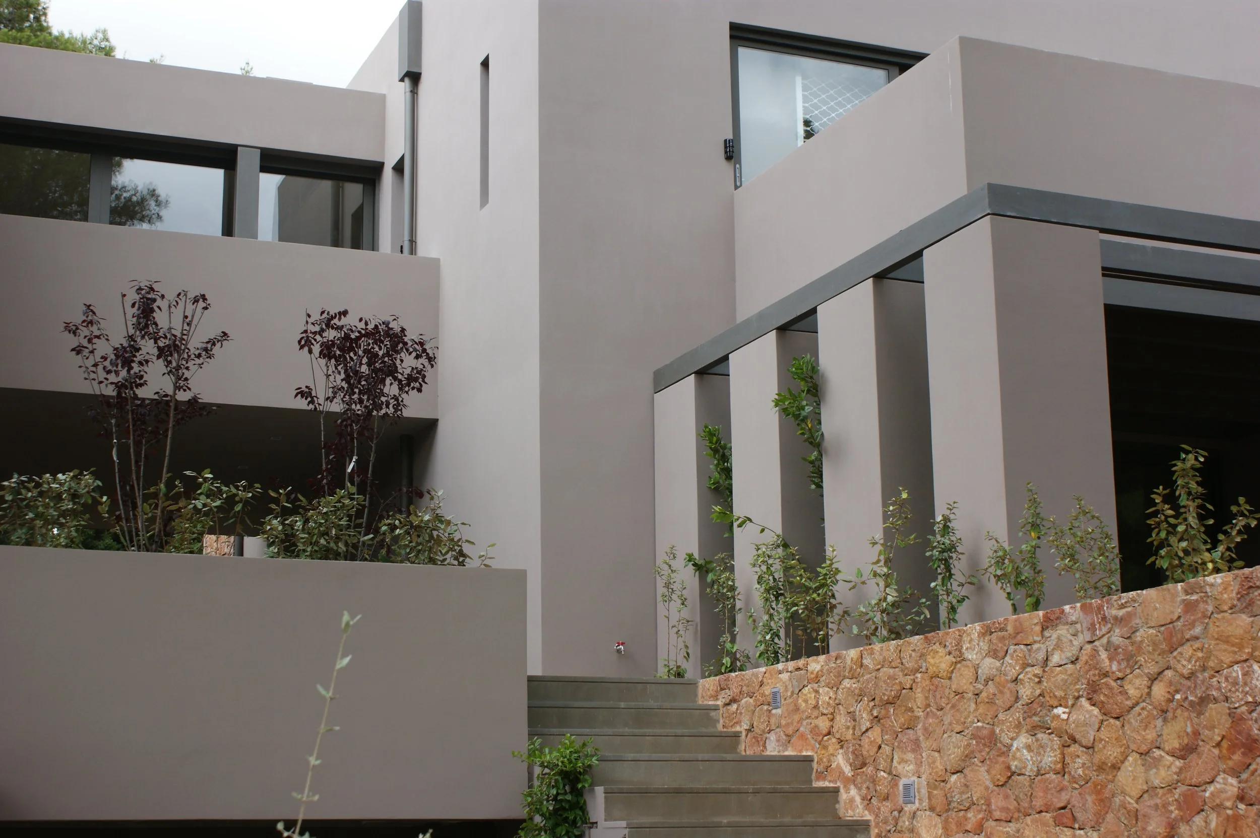 Modern residential building with beige walls, large windows, exterior staircase, and landscaped front yard with bushes and rock wall