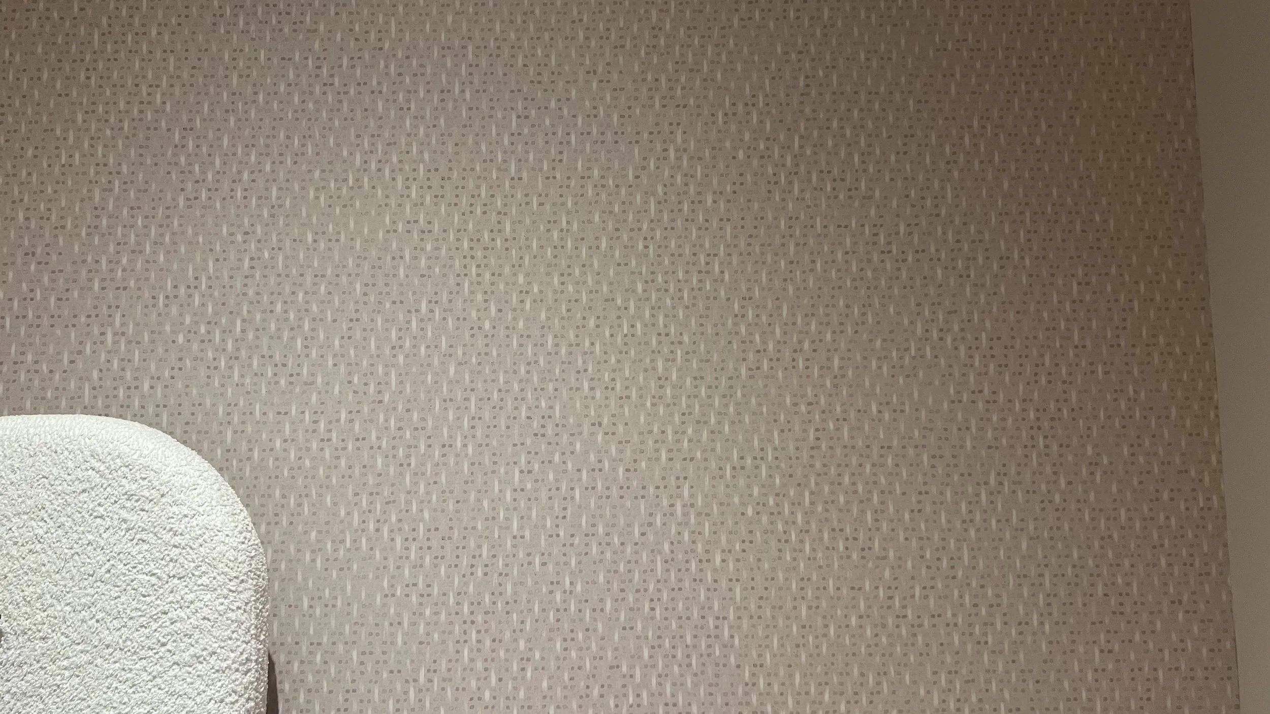 Close-up view of patterned beige wallpaper with small geometric designs and a part of a white fluffy towel in the lower left corner.