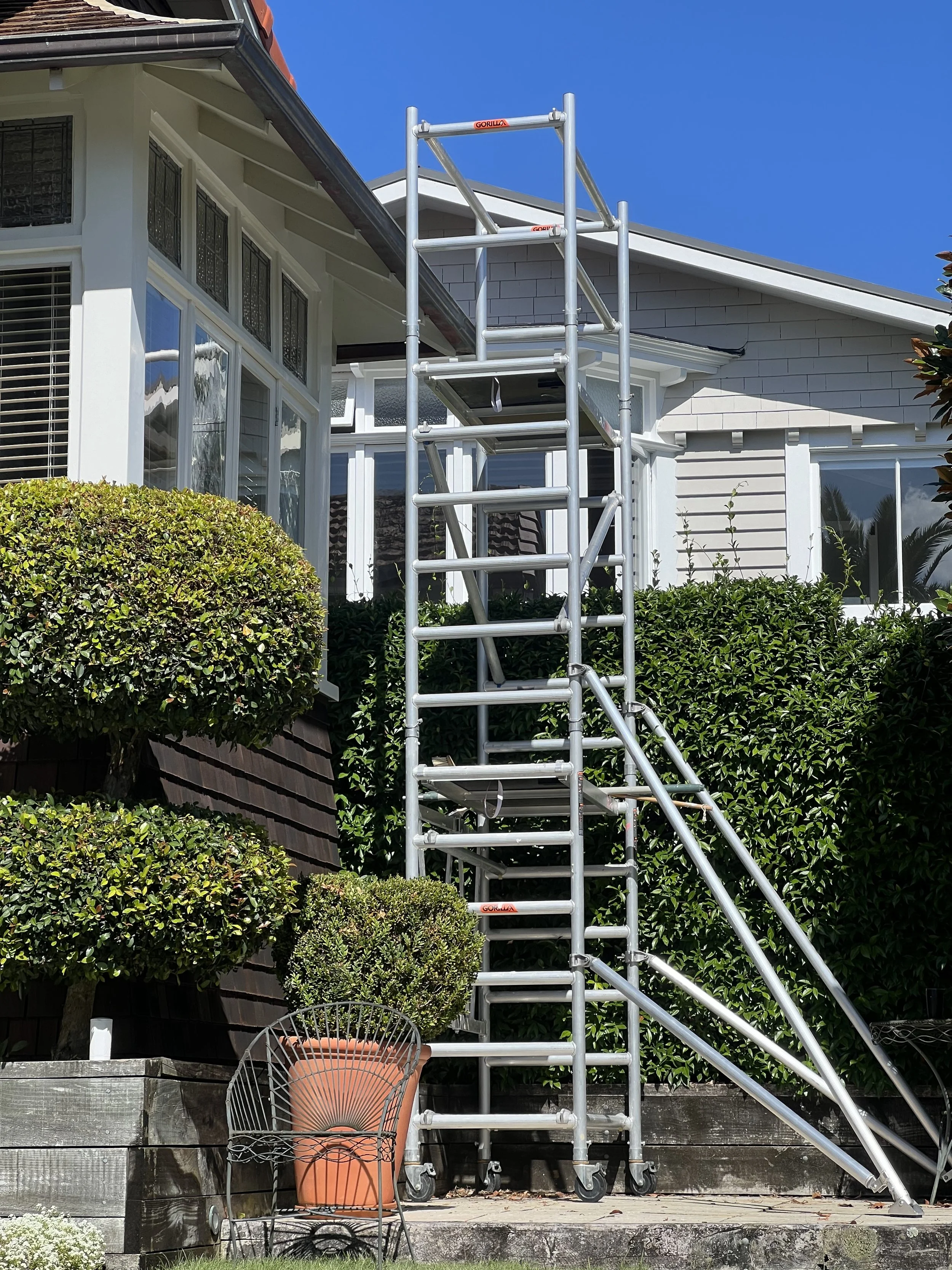 A metal scaffold positioned in front of a house with white siding and large windows. There are green bushes and a potted plant on a wooden planter box in the foreground. The sky is clear and blue.