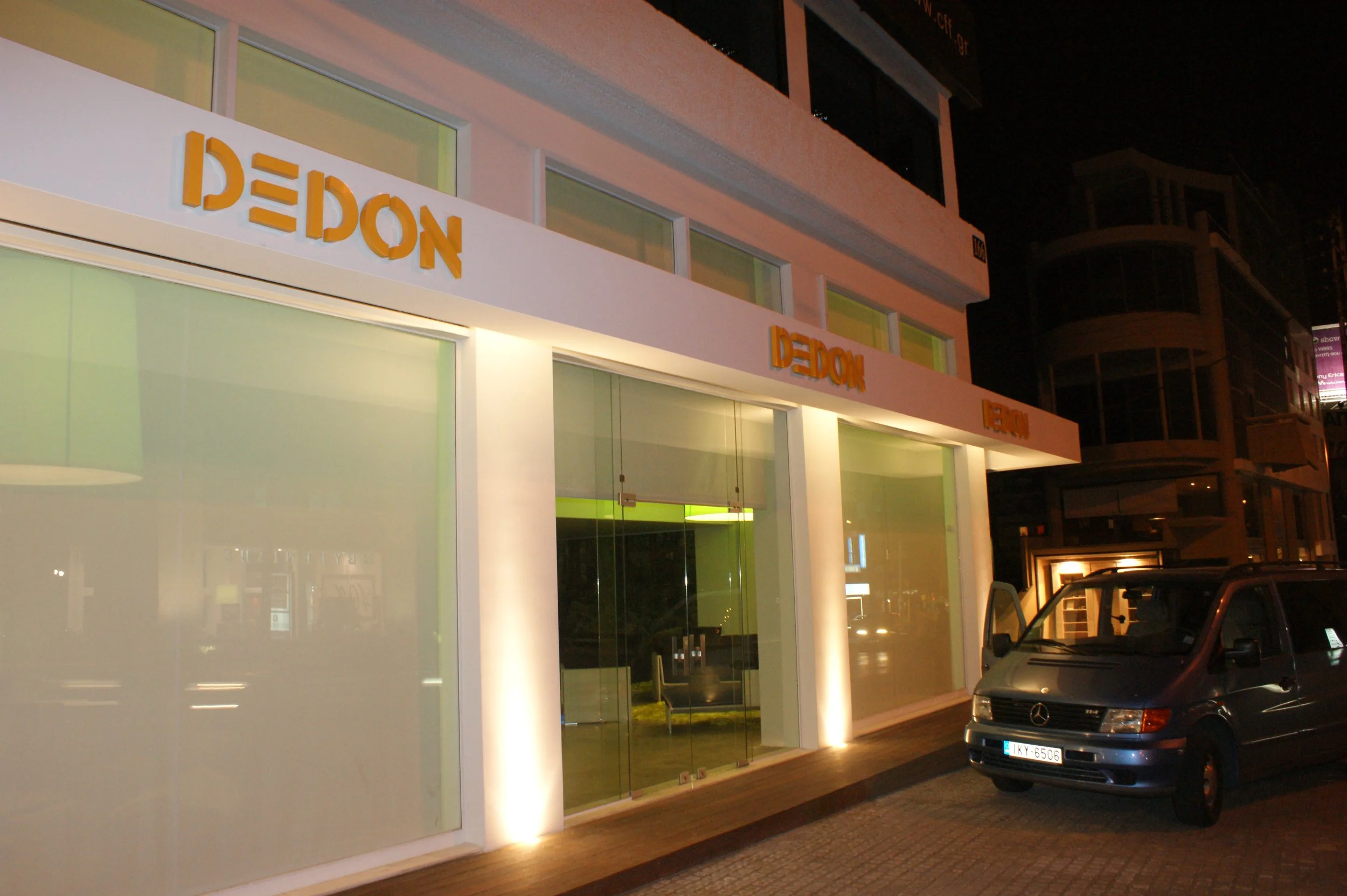 Nighttime view of a modern building with large glass windows and orange 3D letter signage reading 'DE DON' on the upper facade. The building has a white exterior with vertical lighting accents at the lower part, and there are parked cars nearby.