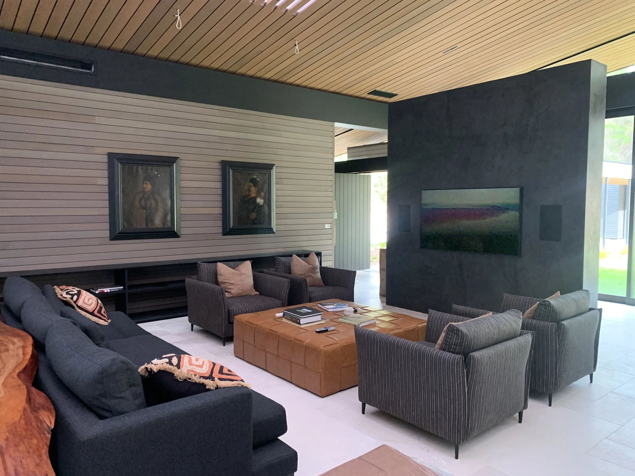 Interior view of a modern living room with hanging light fixtures, wooden wall and ceiling panels, a flat-screen TV on a dark wall, and various sofas and chairs with pillows.