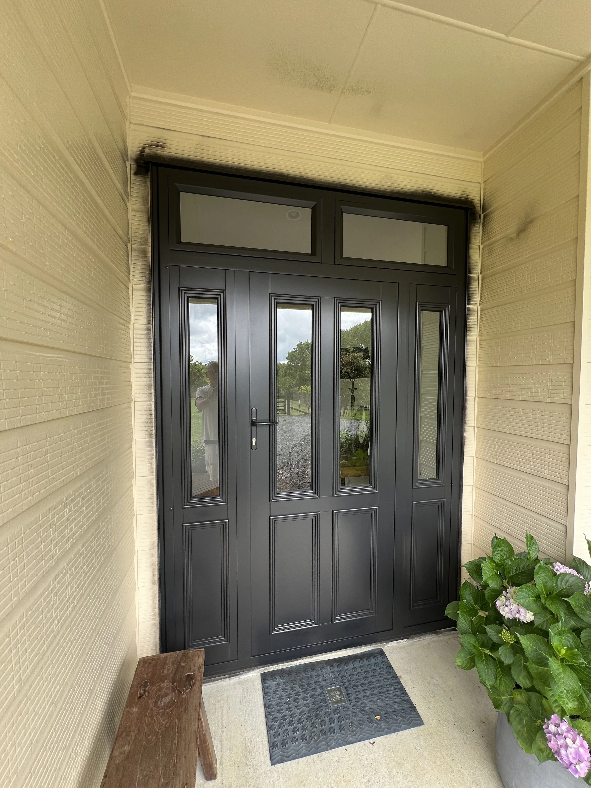 Front porch area with a black double door, beige walls, potted hydrangeas, a brown wooden bench, and a black utility mat on the floor.