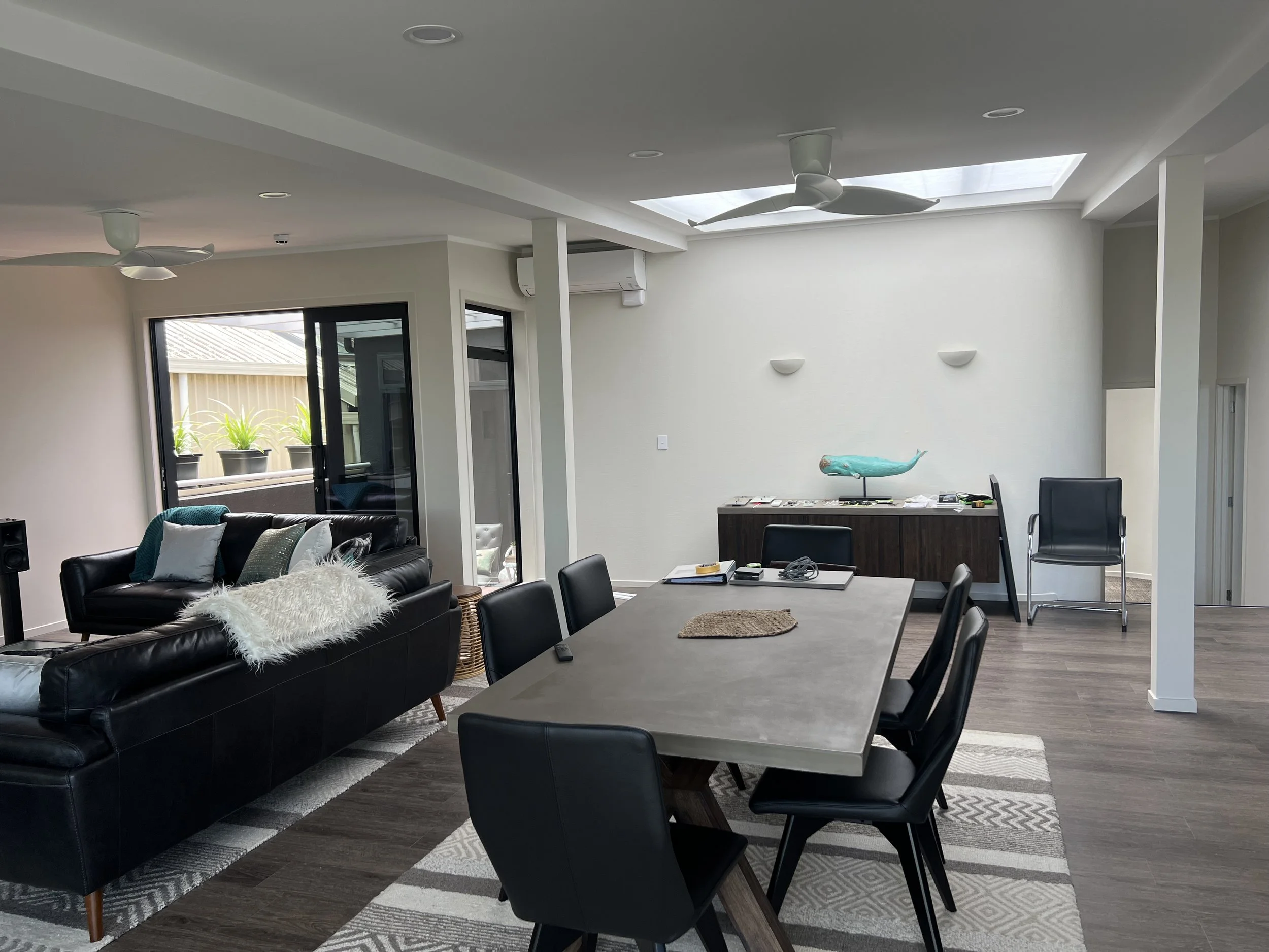 Open-concept living and dining area with a black leather sofa decorated with white throw pillows and a fuzzy throw blanket, a rectangular dining table surrounded by black chairs, a ceiling with two fans and a skylight, large sliding doors leading to 