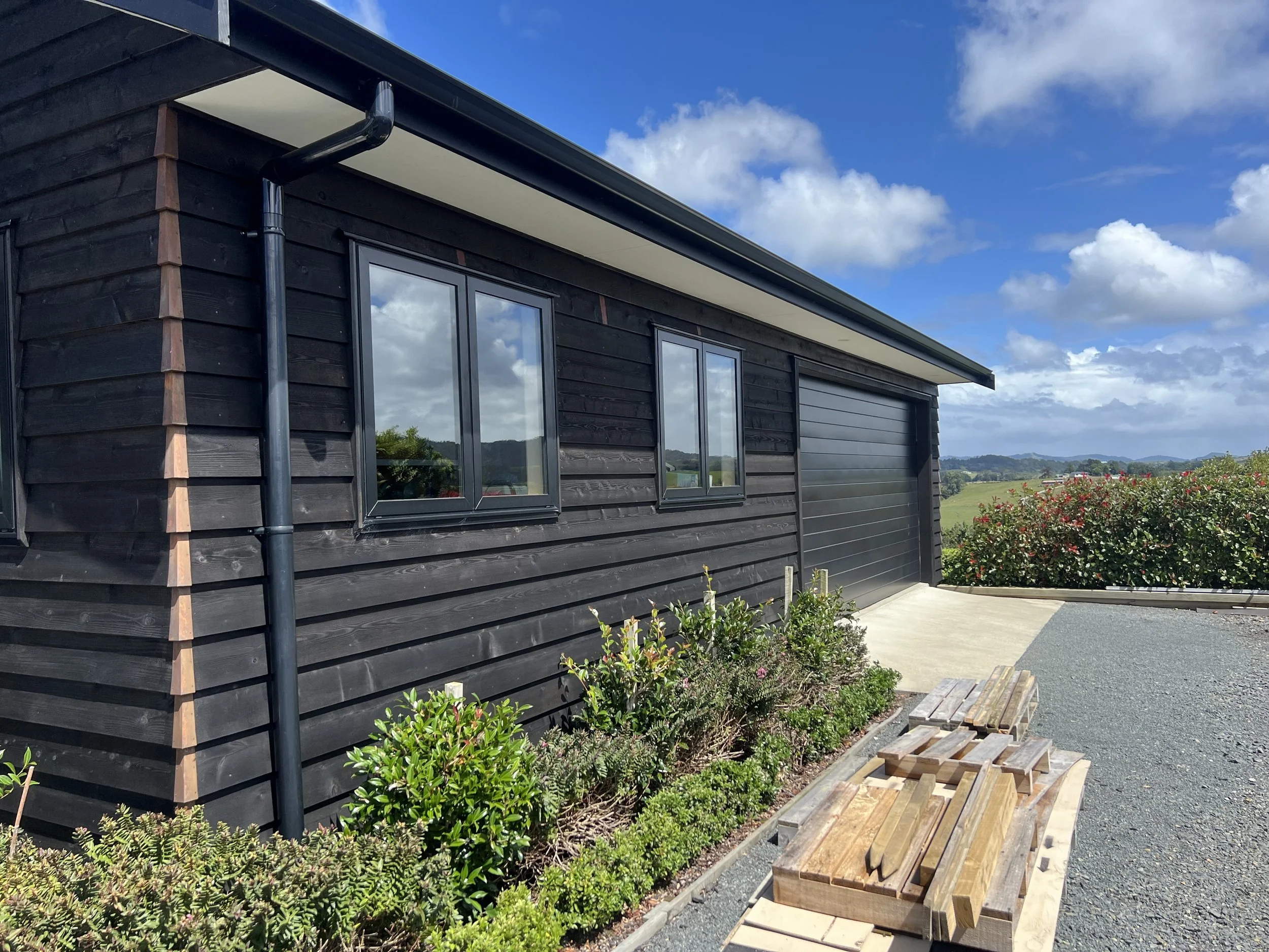 A modern black wooden house with large windows, a black gutter, and a black rain pipe. The house is set in a rural landscape with green fields and distant hills under a partly cloudy blue sky. There is a wooden bench and some stacked wood outside nex