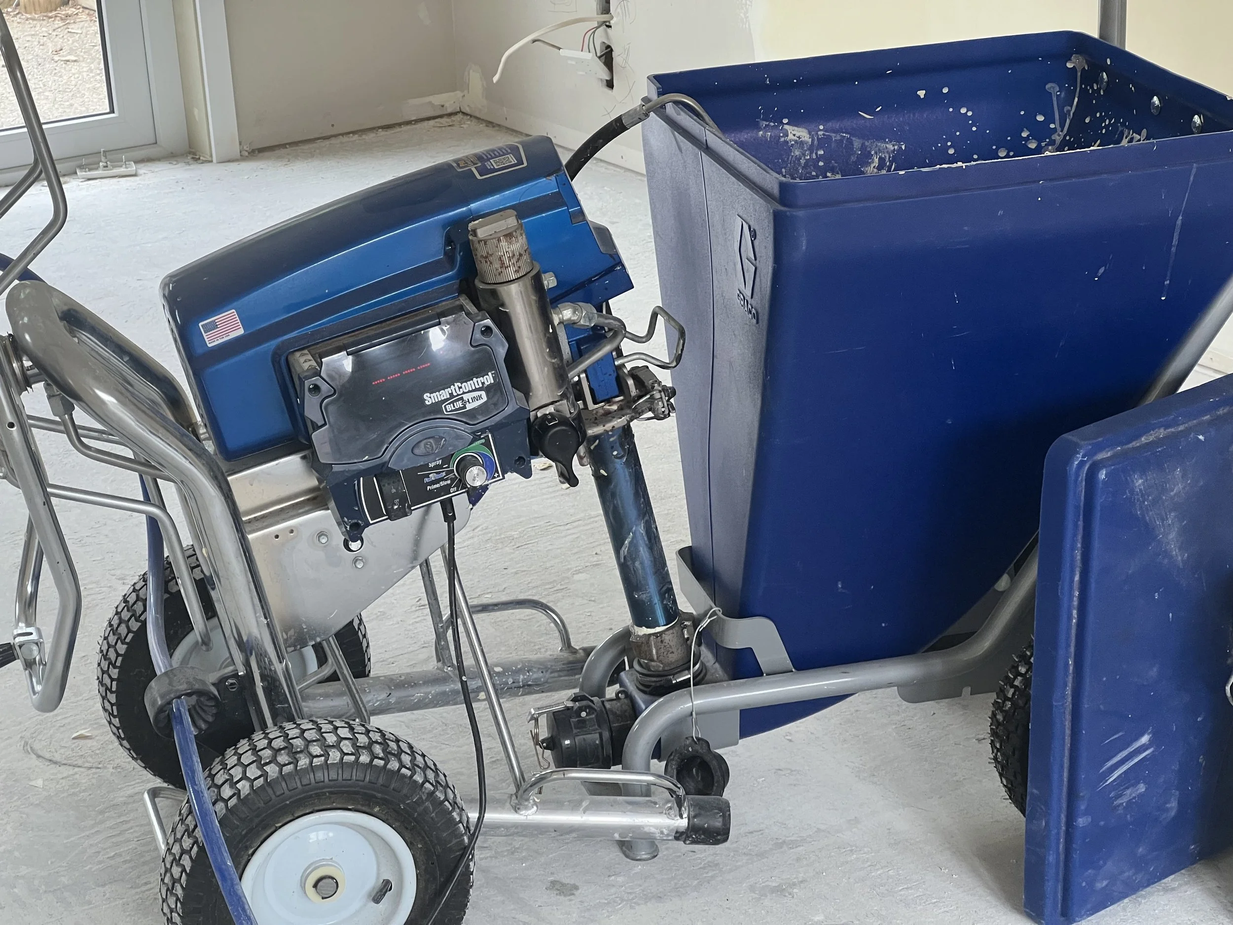 A hospital or medical equipment room with a blue wheelchair and a blue medical cart. The wheelchair has a control panel with various buttons and a joystick, and the cart appears to have splashes of white paint inside.