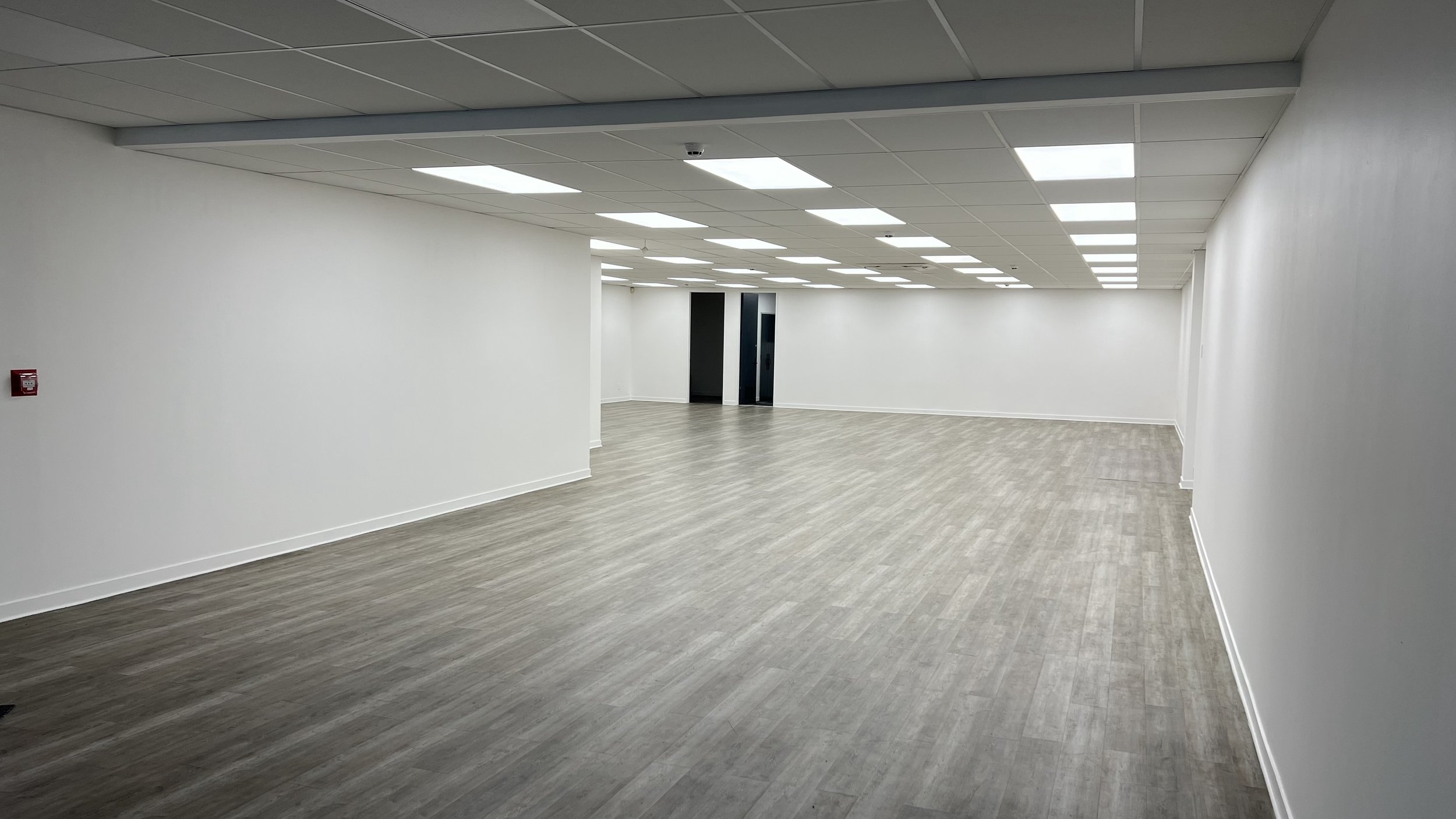 Empty hallway in a modern building with white walls, a ceiling with fluorescent lights, and light wood flooring.