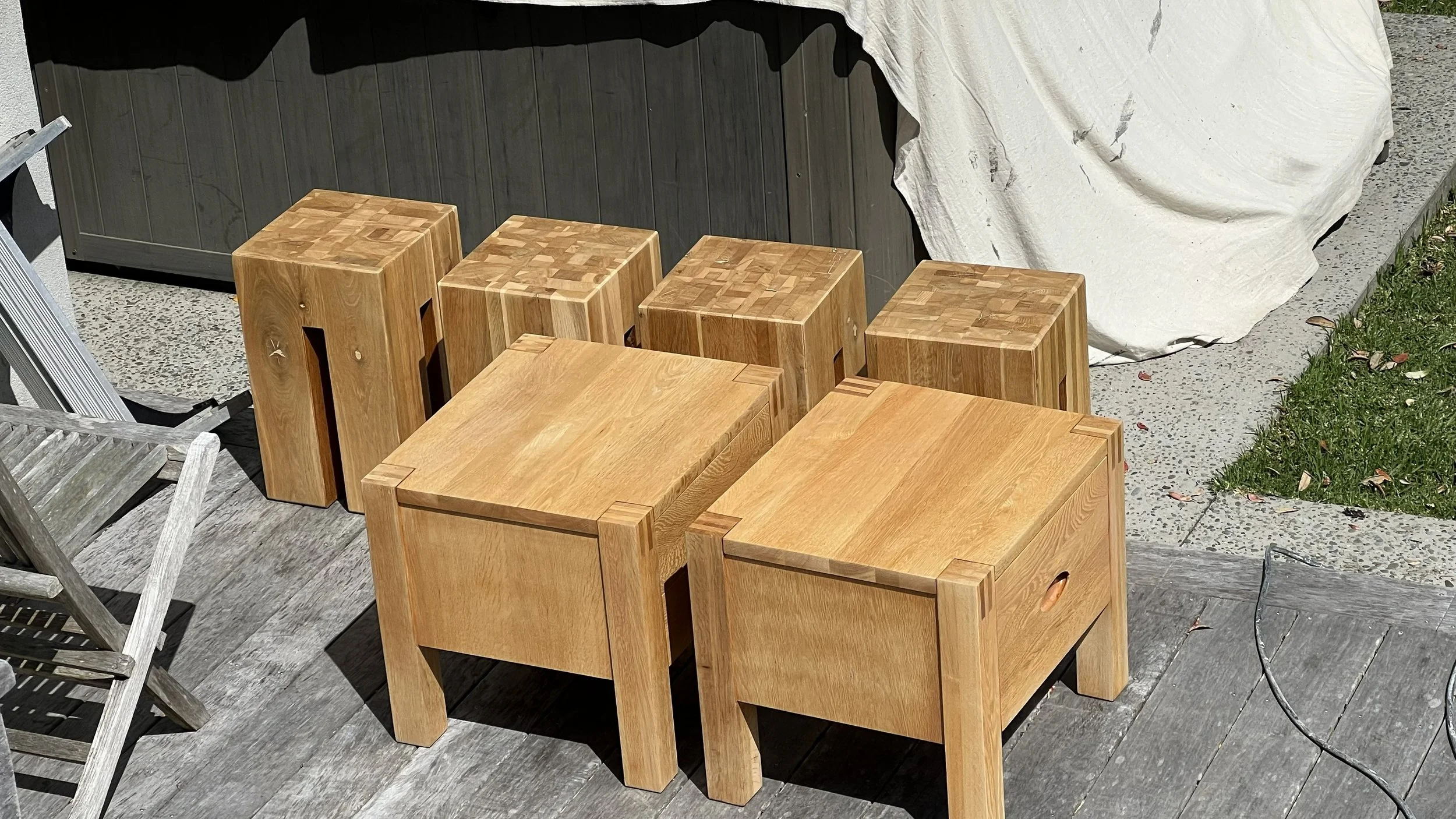 Set of six wooden cubes and two wooden tables on a patio with a dark panel wall, a white tarp, and some grass.