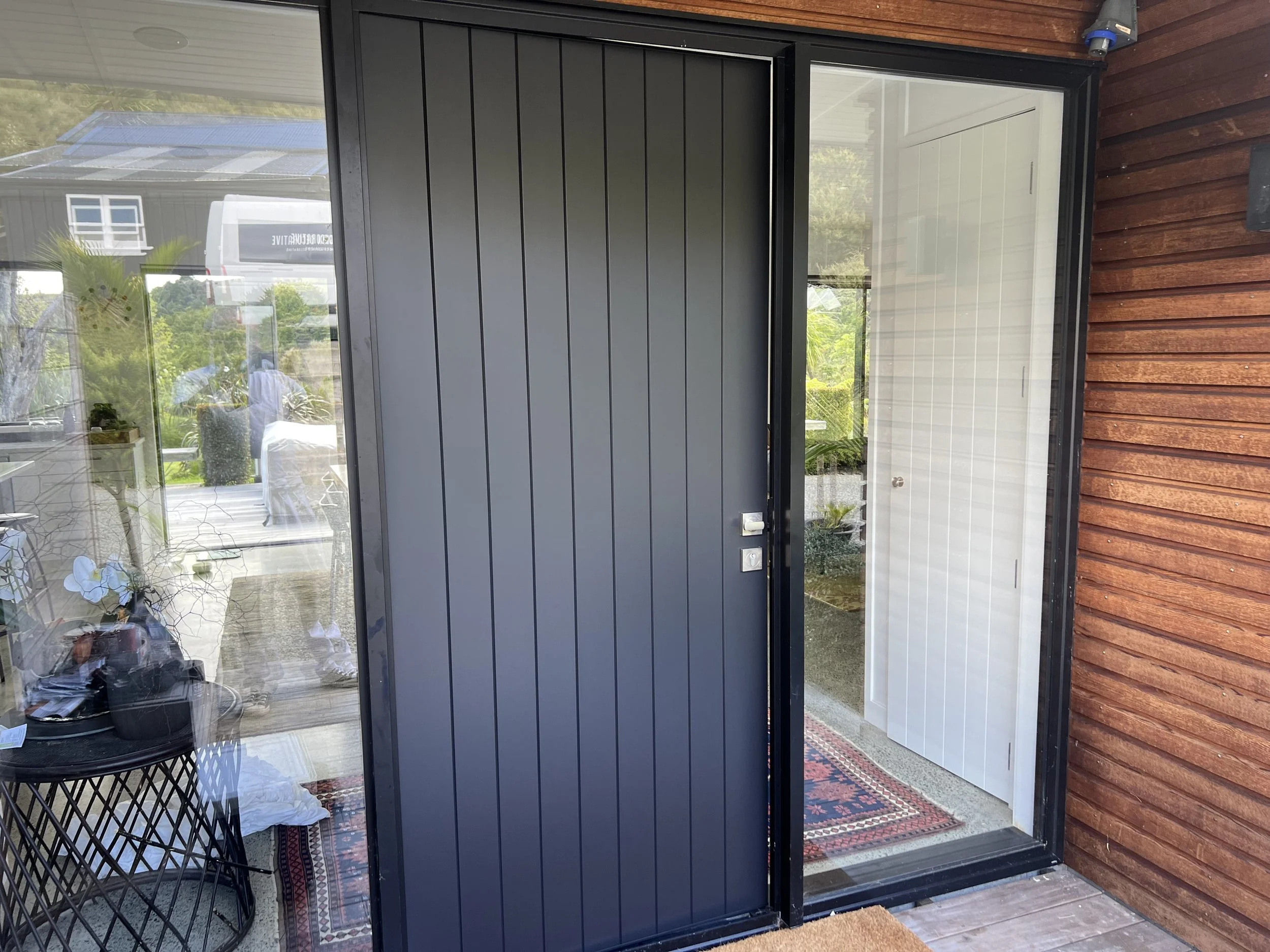 Front view of a modern house entrance with a sliding glass door, dark gray vertical siding, and a white interior door behind the glass. The exterior features wood siding and a decorative red and black rug inside.