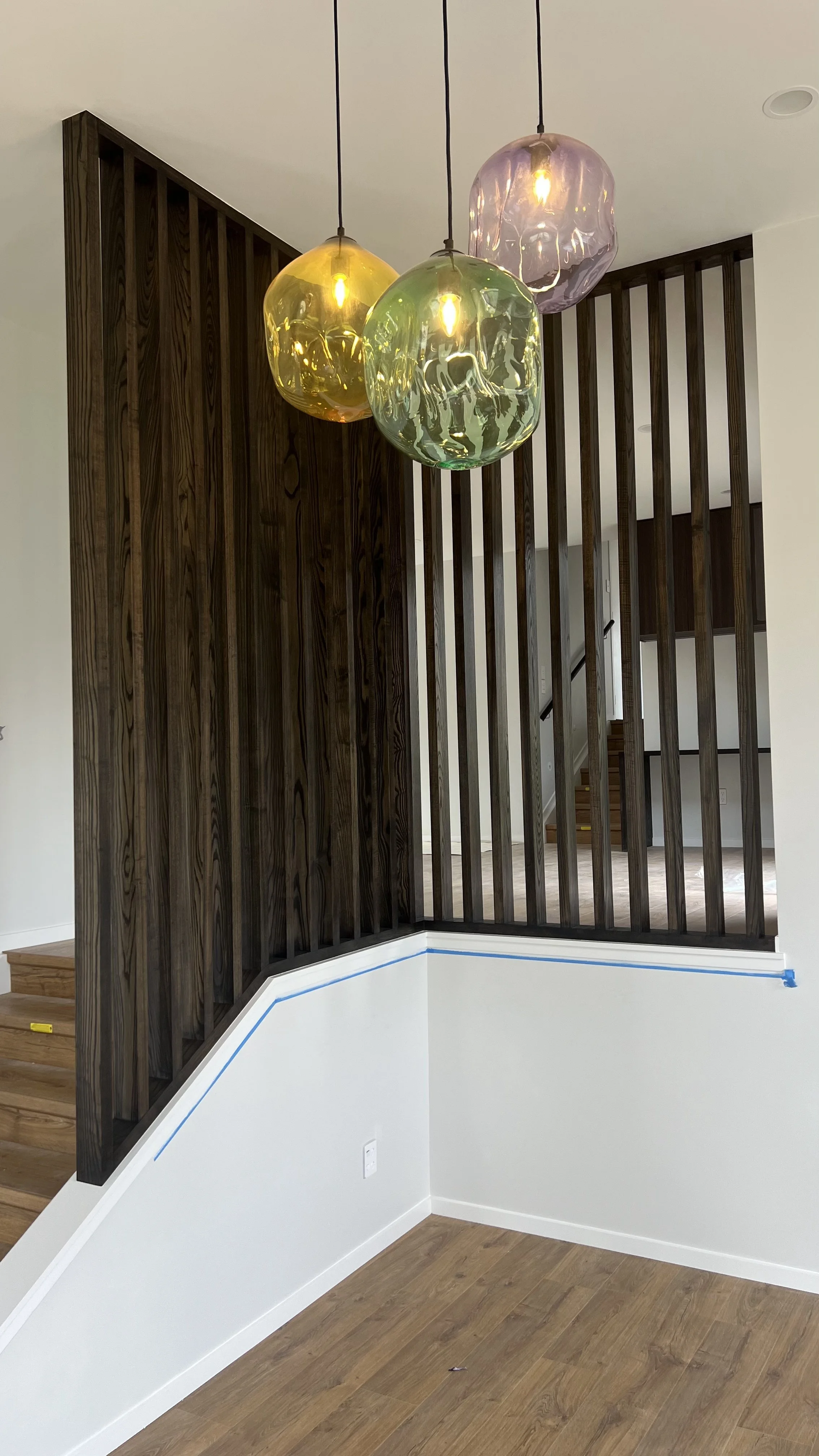 Interior view of a modern home with hanging glass pendant lights, wooden staircase, and a diagonal white wall with blue painter's tape, hardwood floors, and a dark wooden slat room divider.