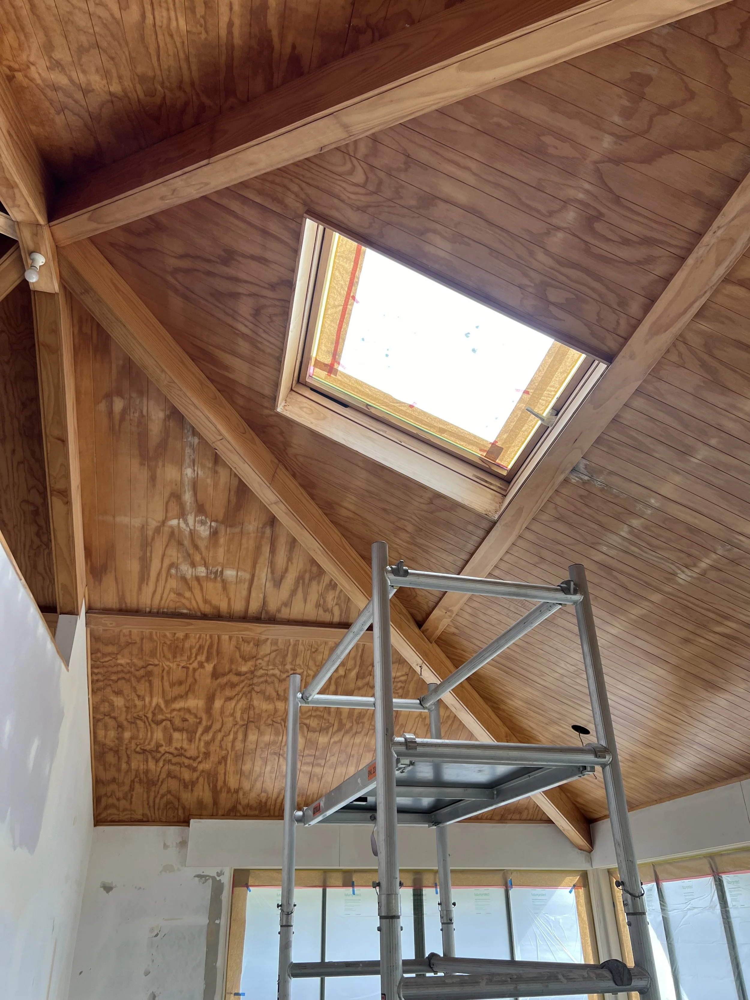 Interior view of a room under construction with wooden ceiling and a skylight, a metal scaffold in the foreground, and windows with translucent coverings.