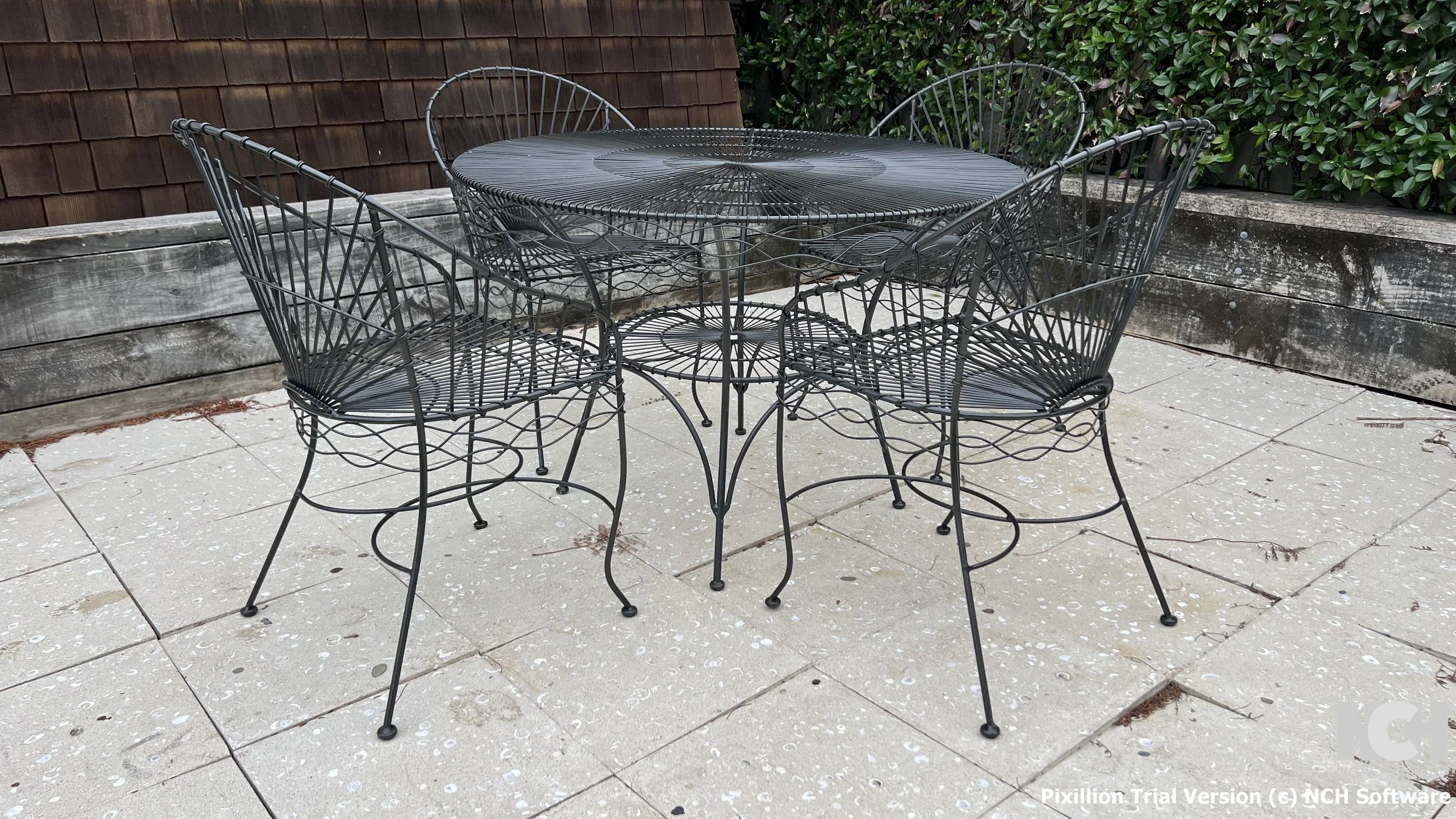 Black metal outdoor dining table with four matching chairs on a patio with concrete tiles, wooden and leafy background.