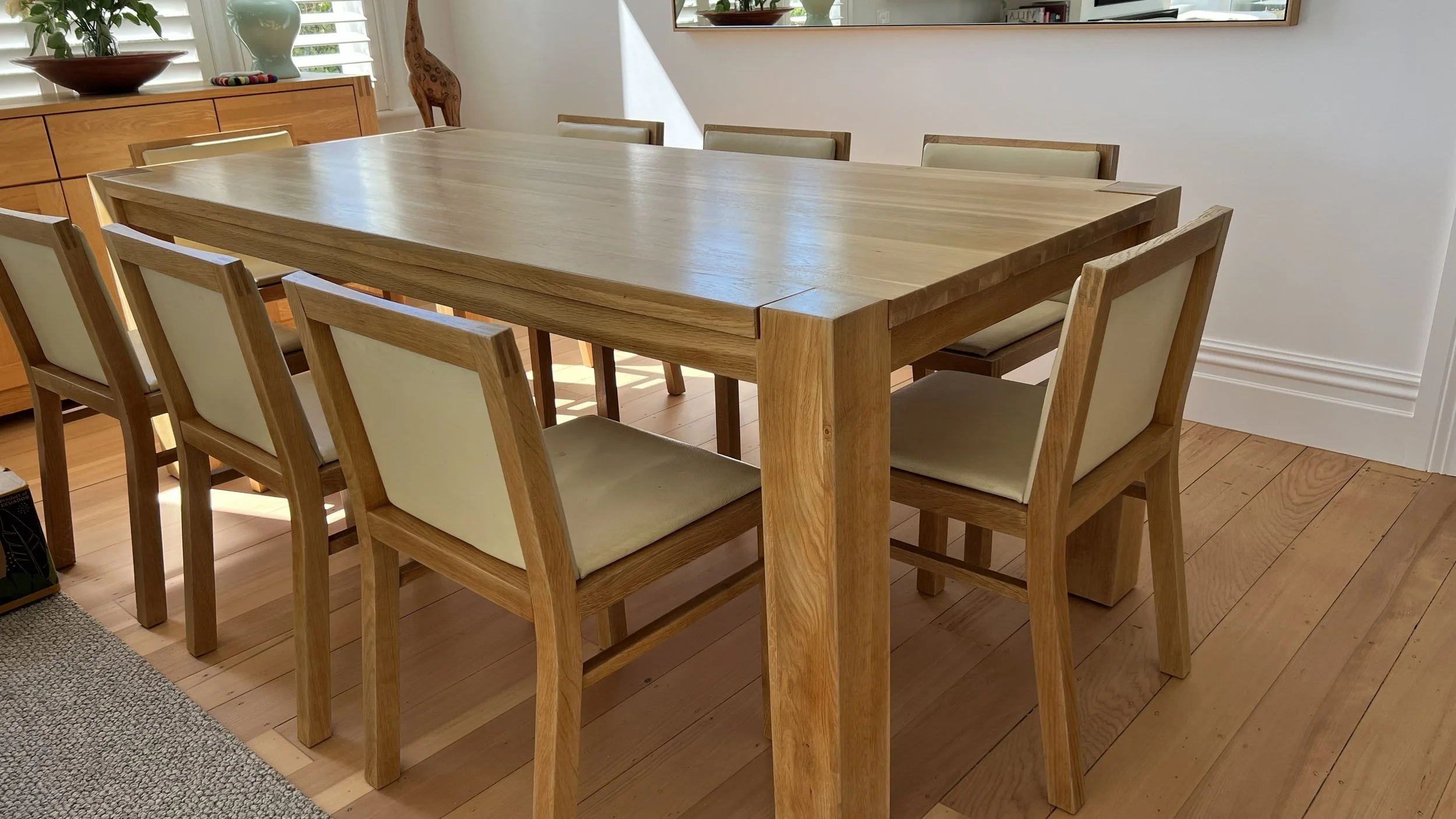 A freshly renovated wooden dining table with eight chairs in a well-lit room, with a sideboard and decorative items in the background.