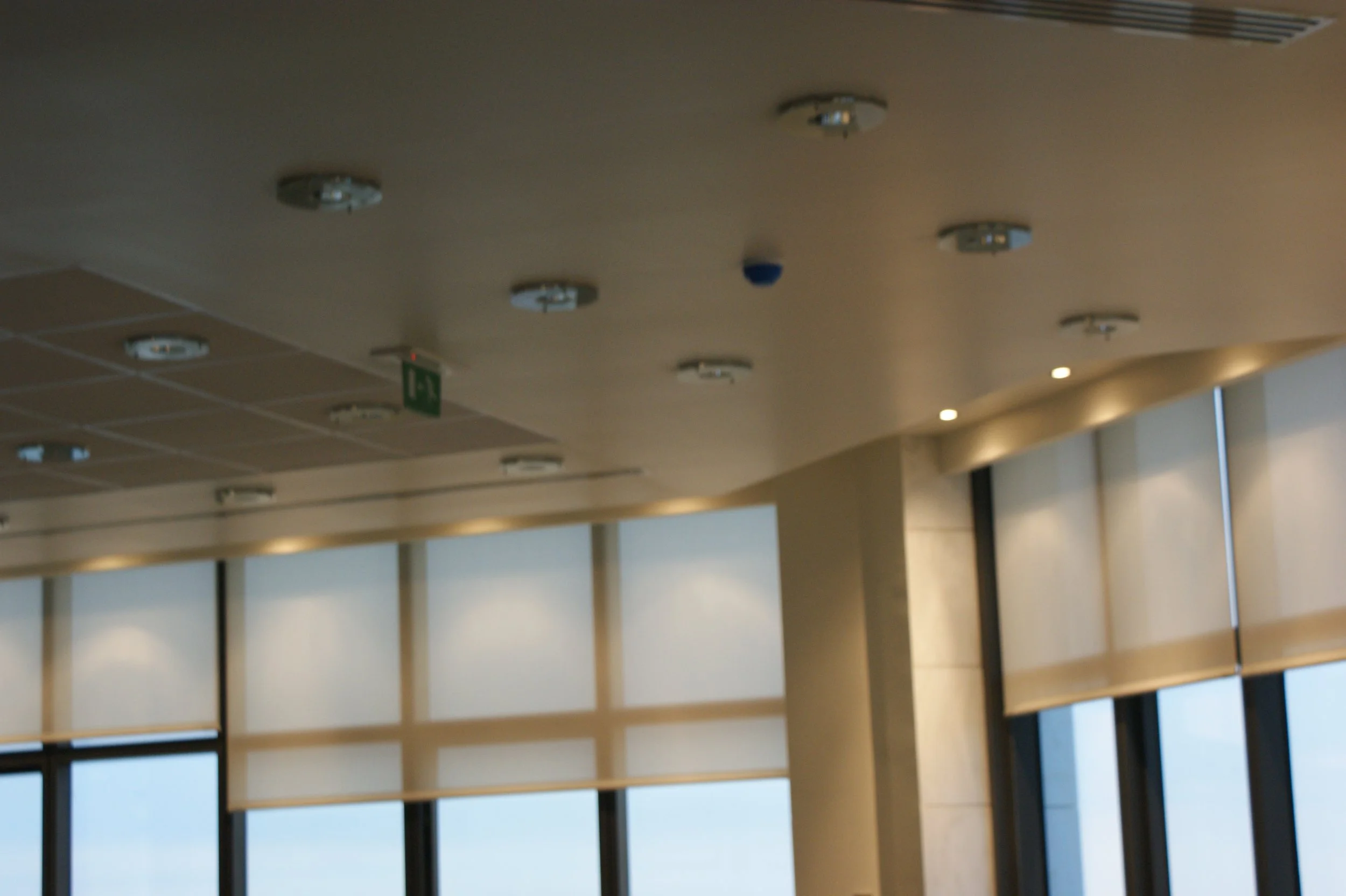 Ceiling with several fire alarm and smoke detector devices, outside light, and an emergency exit sign in a modern interior space.