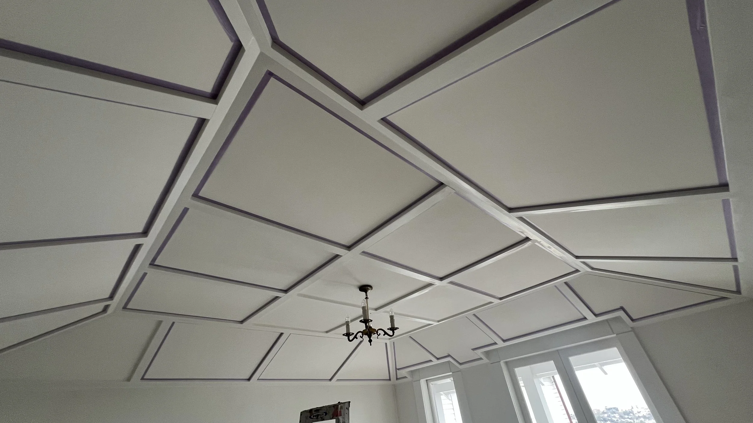 Ceiling with geometric pattern design and a chandelier hanging in the center, with windows on the side allowing natural light.
