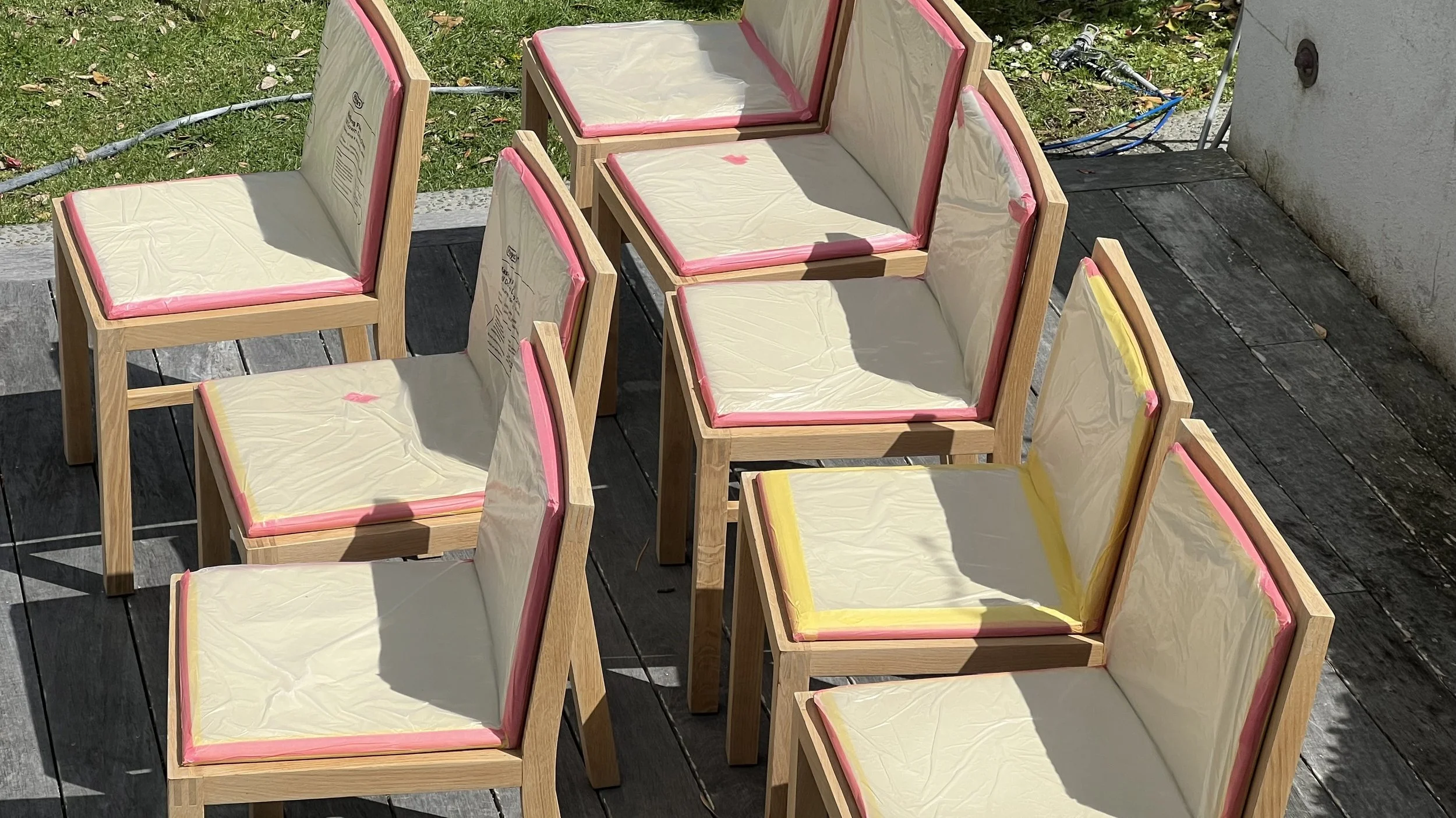 Outdoor scene with several wooden chairs covered in plastic, arranged on a wooden deck with grass and leaves in the background.