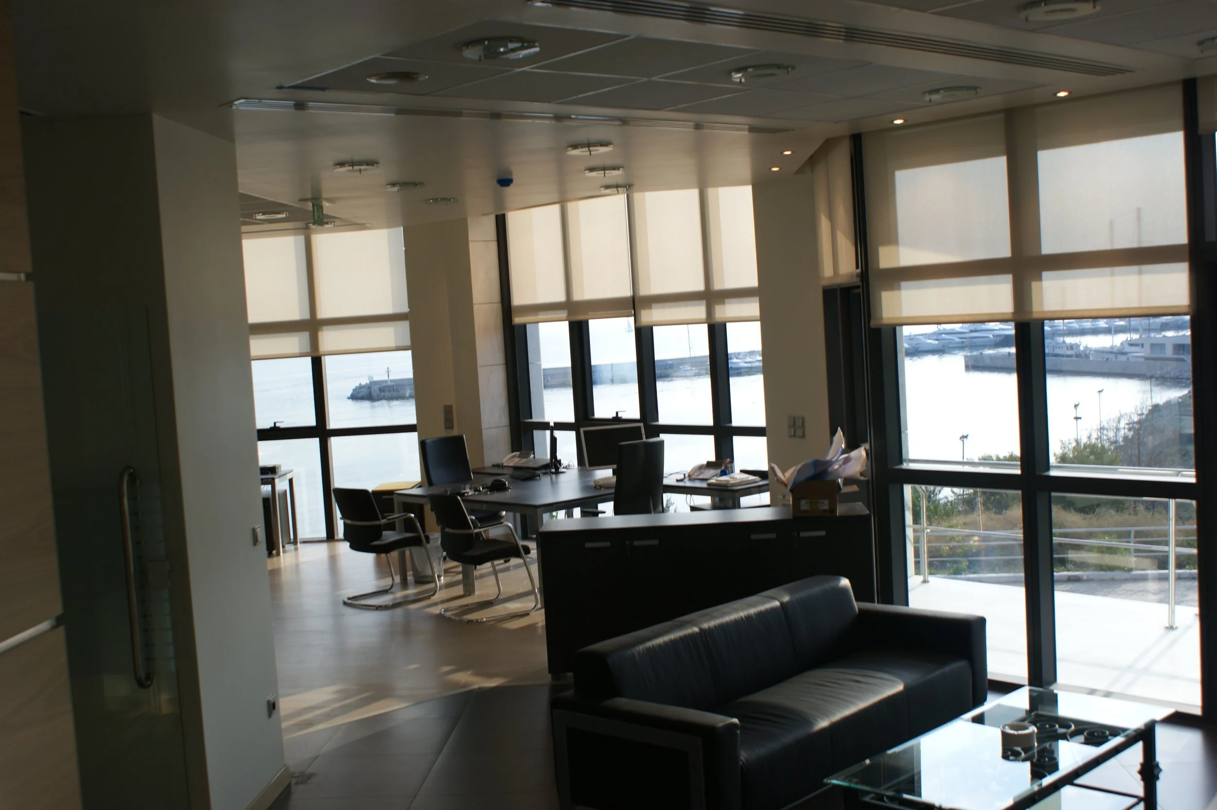 An office with large windows overlooking a waterfront with boats, a conference table with chairs, and a black sofa.