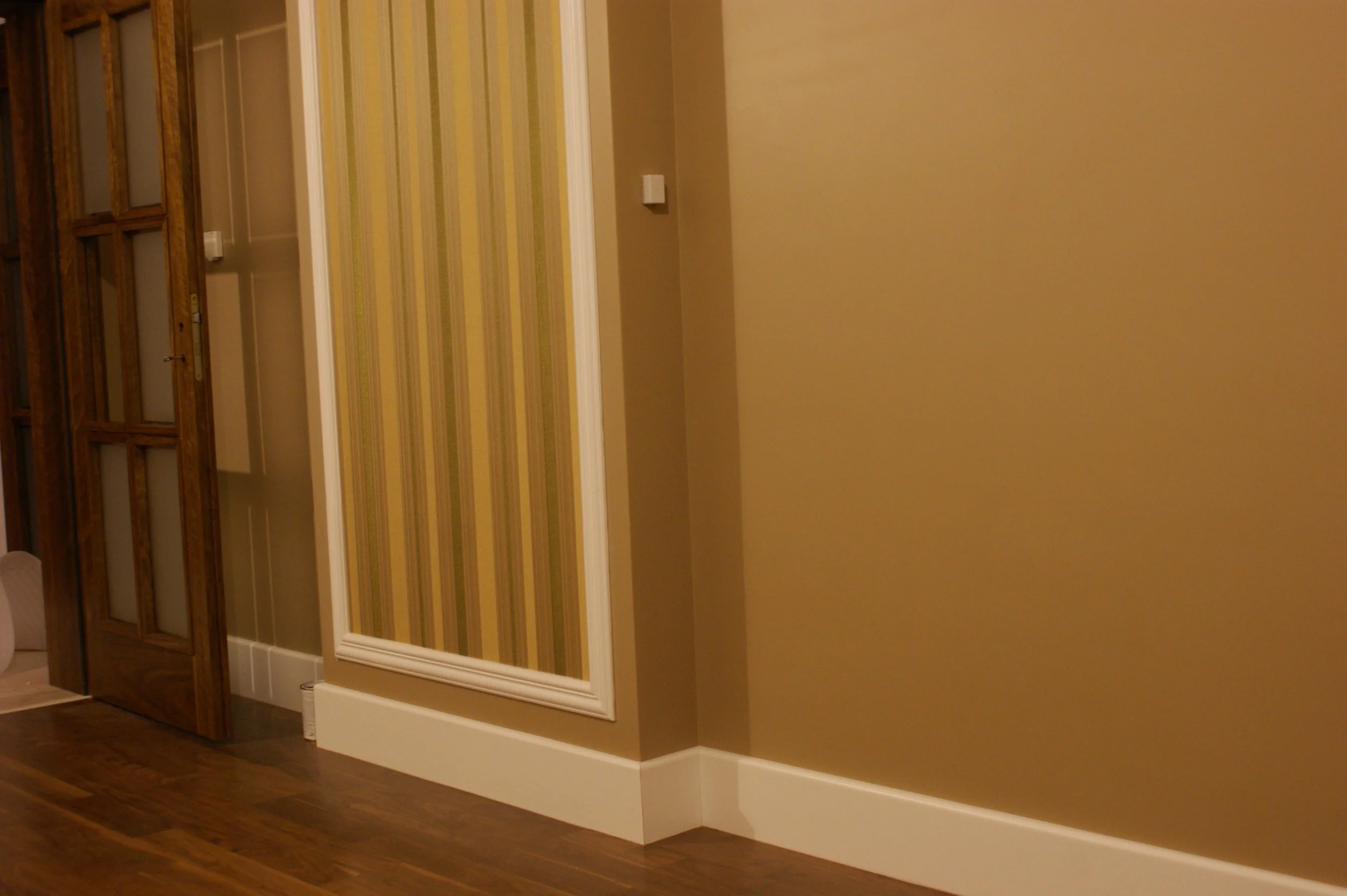 Interior of a house showing a wall with beige and green striped wallpaper, a white picture frame, and a thermostat on the wall. Part of a wooden door with glass panels and the wooden floor are visible.