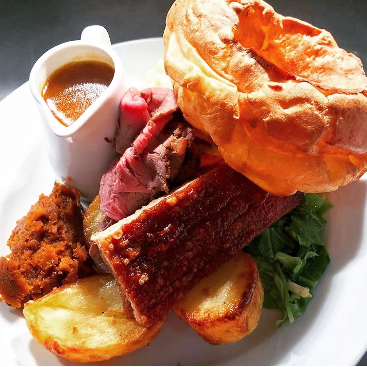 A plate of traditional British Sunday roast with roast beef, Yorkshire pudding, roast potatoes, gravy, and vegetables, served with a small pitcher of gravy.