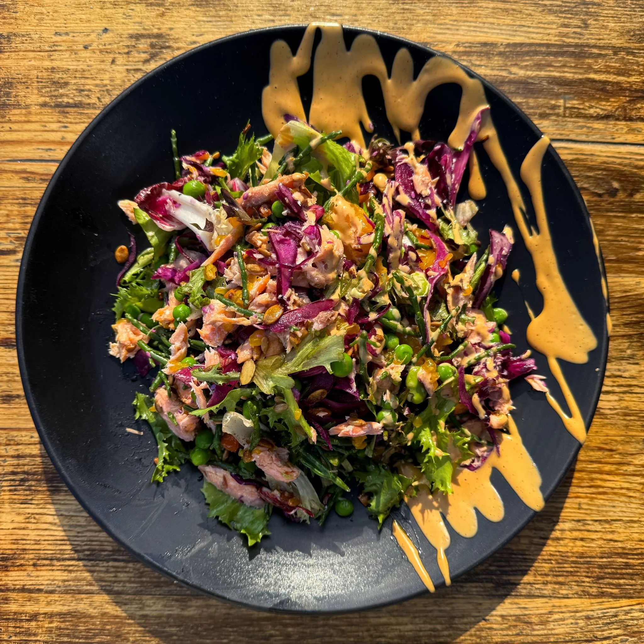 A black plate with a mixed green salad topped with a dressing and garnished with nuts and seeds, on a wooden table.