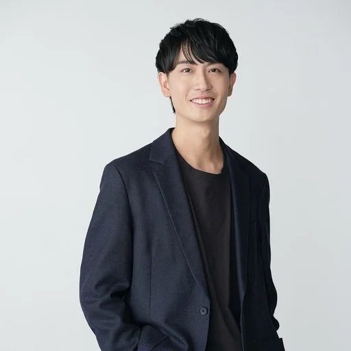 黒いシャツとダークブルーのジャケットを着た若い男性が白背景の前で笑顔で立っている写真