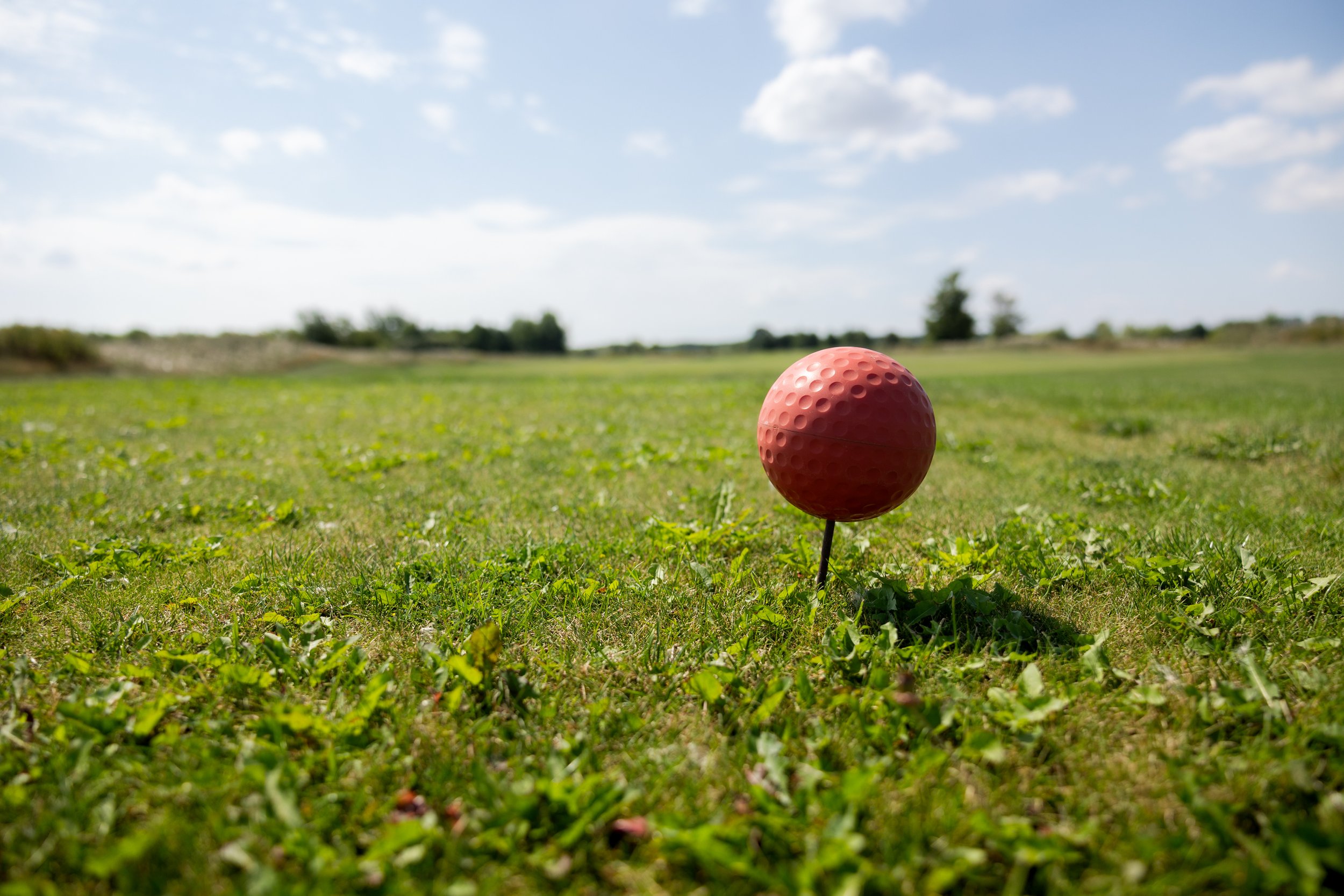Roter Golfball auf einem Golfplatz mit grünem Rasen. Im Hintergrund sonniger Himmel mit Wolken und Bäume.