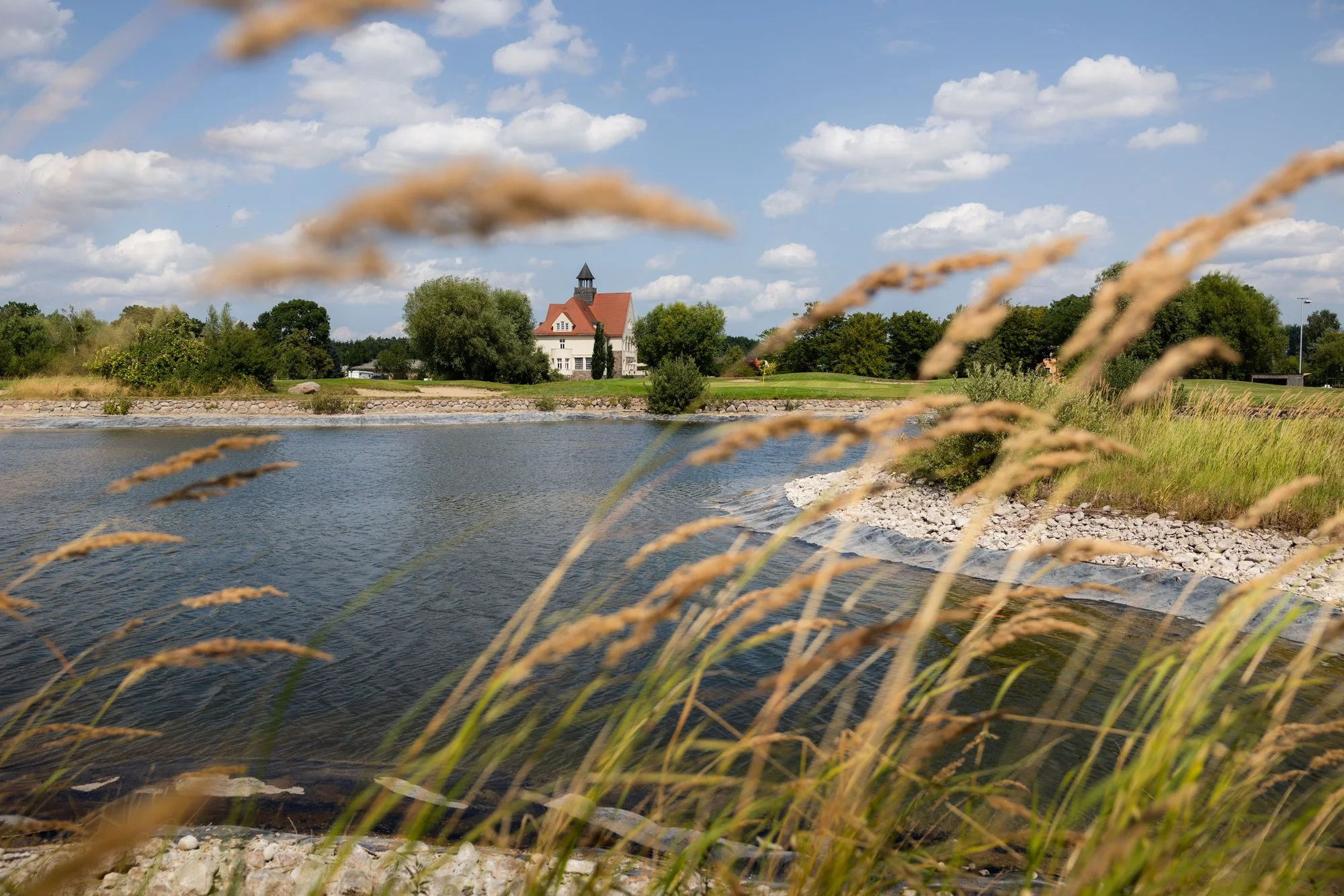 Schloss umgeben von grünen Bäumen, am Ufer eines Teichs bei sonnigem Himmel mit Wolken.