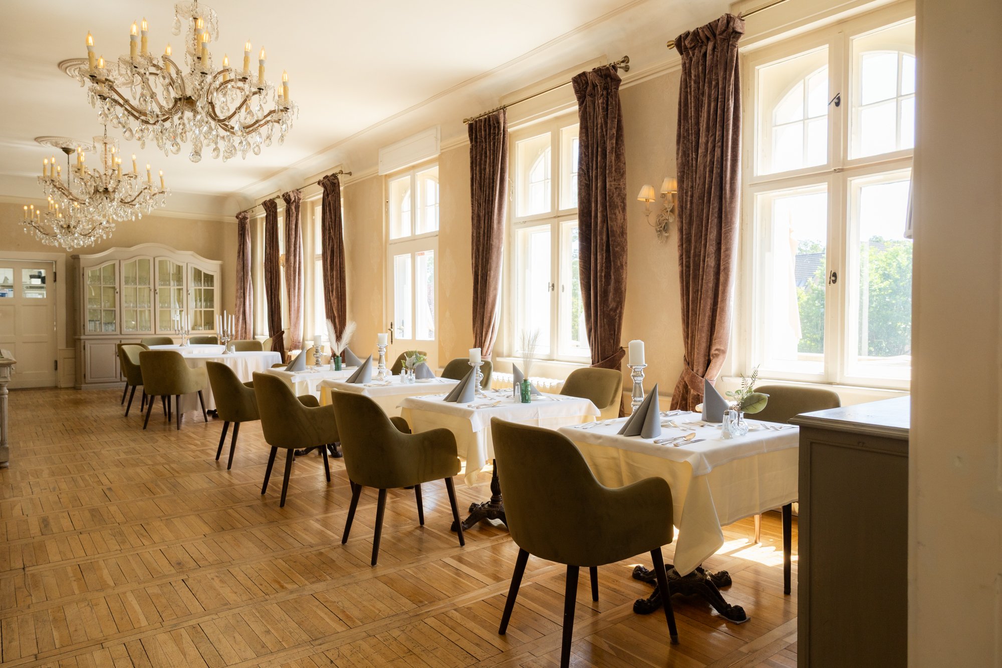 Eleganter Speiseraum im Schloss mit großen Fenstern, schweren Vorhängen, mehreren Tafelrunden mit weißen Tischdecken und Kerzen, und zwei große Kronleuchter an der Decke.