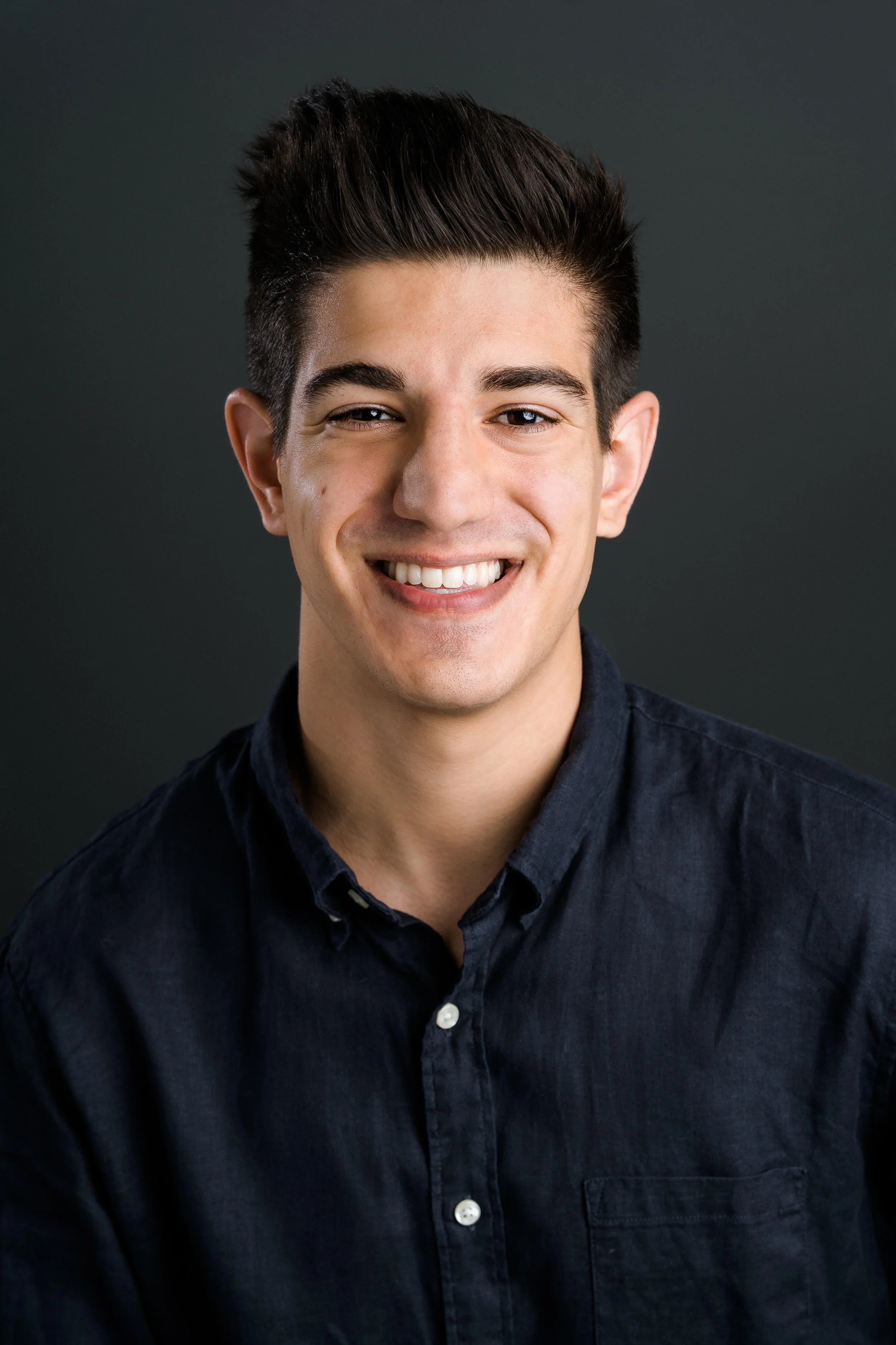 Portrait of a young man with dark hair, smiling, wearing a dark button-up shirt, against a dark background.
